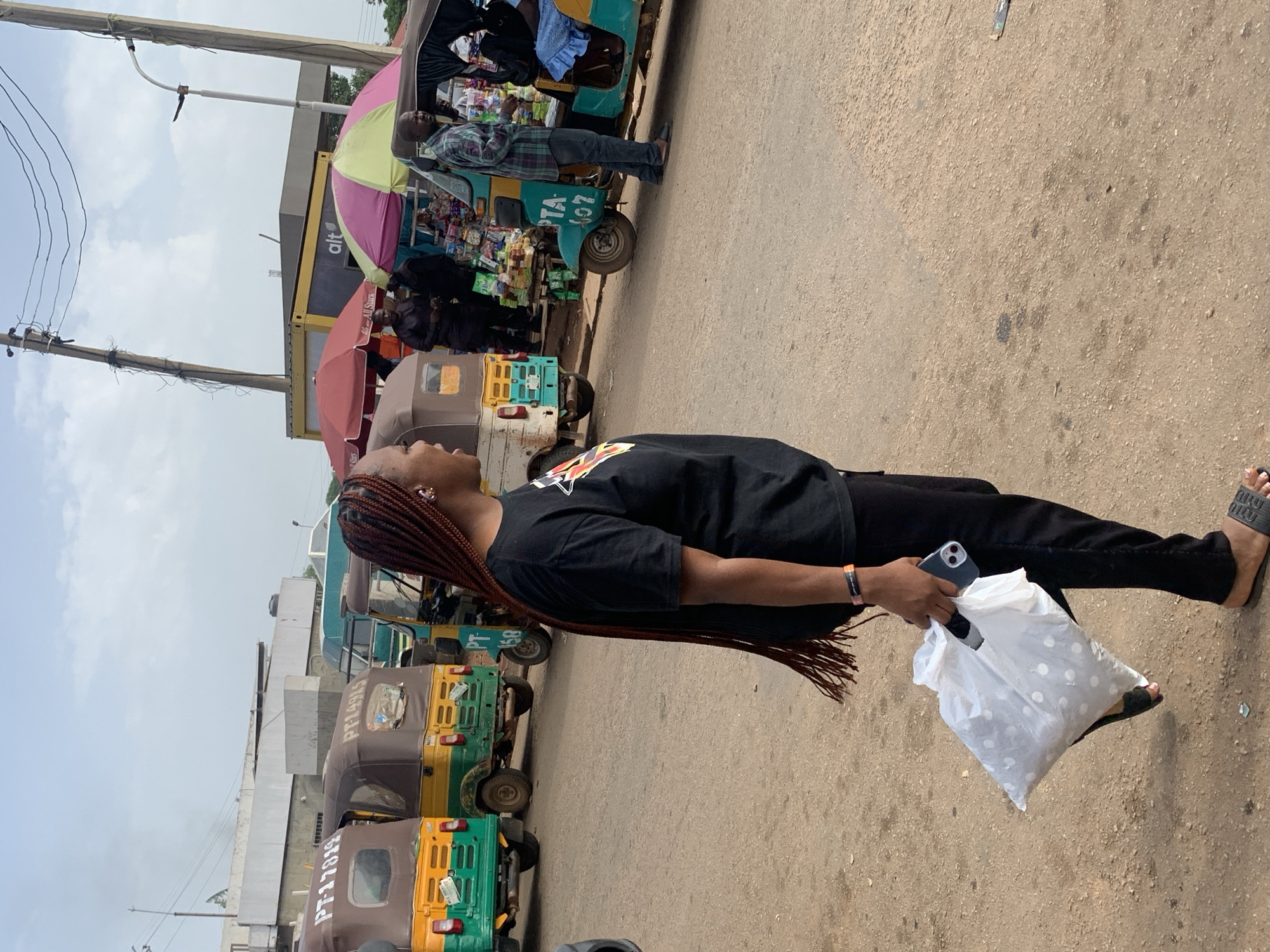 In Gura Top, Nigeria, a woman with long, braided hair walks with a white plastic bag in her left hand and a smartphone in her right hand. She is wearing a black t-shirt and black pants, with black slides on her feet. The scene appears to be a busy street market with several auto-rickshaws (also known as Keke Napeps) parked or driving. Some of the auto-rickshaws have people inside and some are being used as mobile vending stalls with colorful umbrellas. In the background, there are buildings, utility poles with overhead wires, and a cloudy sky. The ground is a dirt and gravel road.
