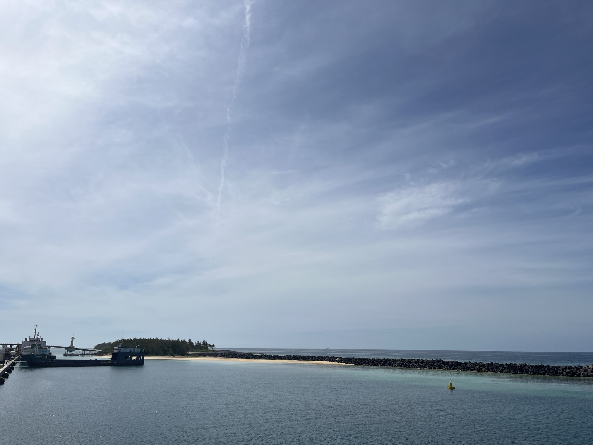 The image depicts a coastal scene in Unknown, Unknown. In the foreground, calm, blue-green water fills the lower portion of the frame, with a small yellow buoy visible slightly right of center. Along the shoreline, a sandy beach is bordered by a rock breakwater extending into the sea. To the left, a structure appears to be a dock or pier complex with industrial-type buildings and a vessel attached. Further in the distance, a small, green island or peninsula is visible. The sky is overcast with scattered clouds, creating a soft light across the scene.