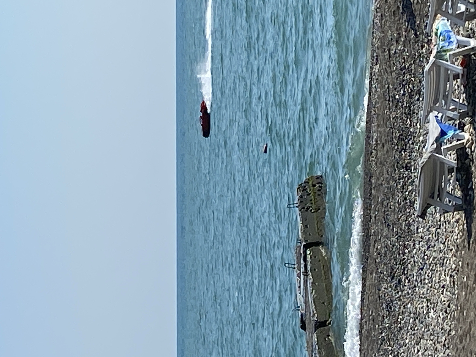 The image shows a beach in Chabkwara, Abkhazia. In the foreground, there are several lounge chairs on a pebble beach. In the middle ground, a concrete pier extends into the sea. The sea is blue with small waves. In the background, a red jet ski is moving across the water, creating a wake. The sky is clear and blue.