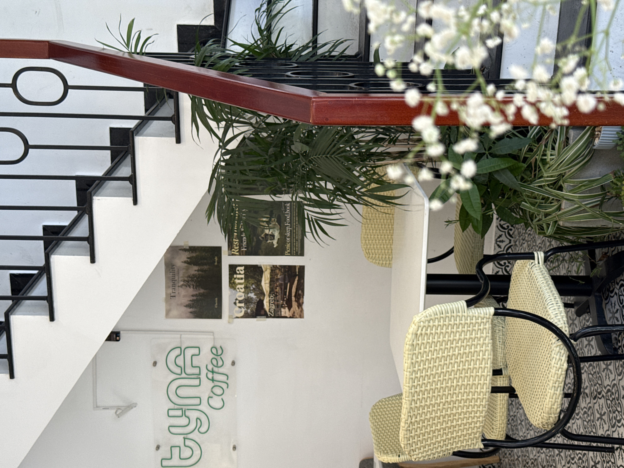 This image shows the interior of a cafe named "Eyna Coffee." Located in Da Lat, Vietnam, the cafe features a staircase with white steps, black railing, and a reddish-brown wooden handrail. Potted plants adorn the staircase and the surrounding area, with some white flowers in focus in the foreground. Posters with images and text such as "Tranquility," "Croatia," "Rest," and "Picnic or sleep, Food, book" are attached to the wall beside the staircase. Two wicker chairs with black metal frames are visible, situated on a patterned tile floor. A neon sign bearing the cafe's name in green letters is fixed to the wall.