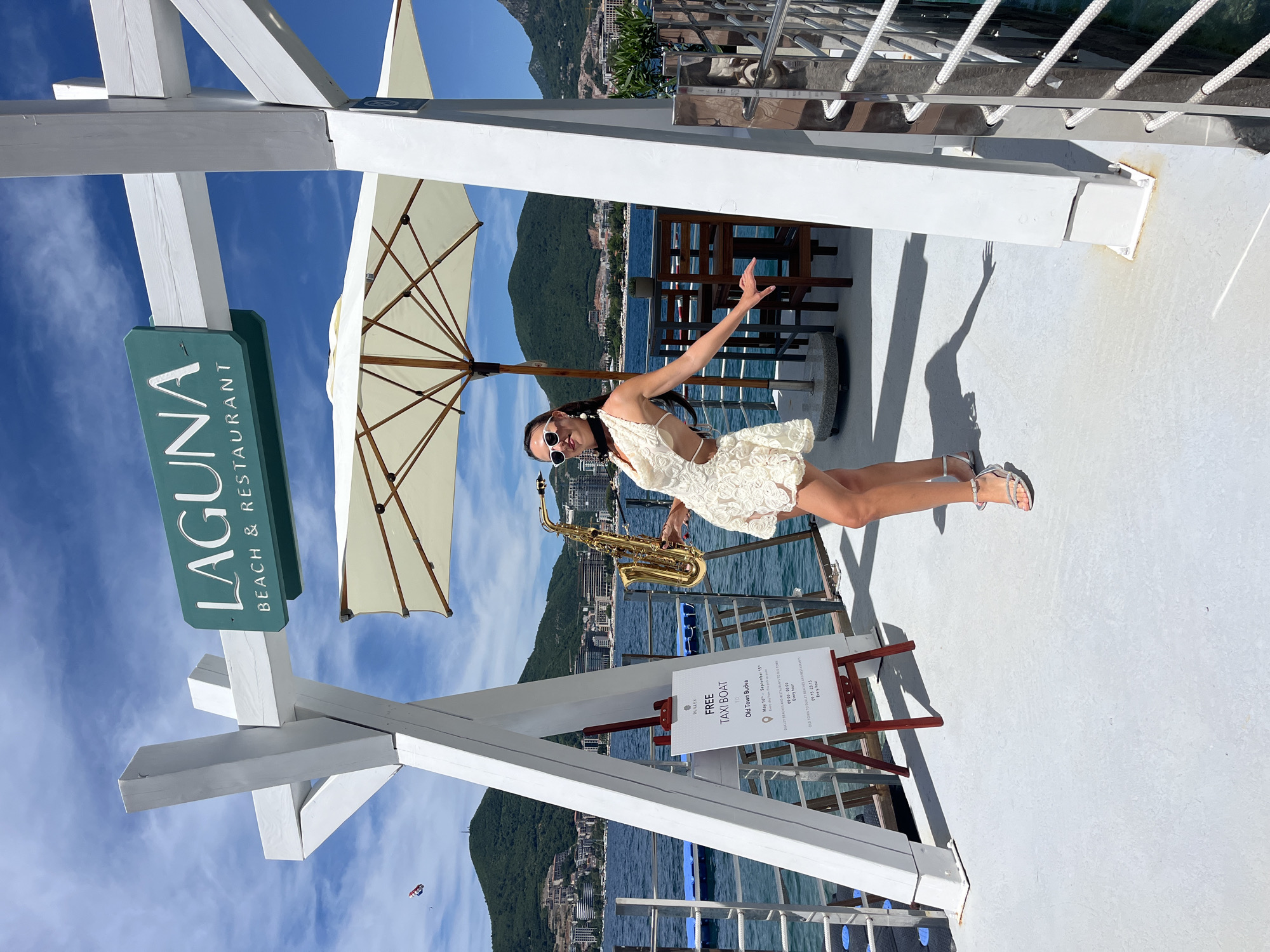 A woman in a short, white, lace dress and silver sandals stands on a white wooden deck, holding a saxophone. She wears sunglasses and a black neck scarf. Behind her, a sign reads "LAGUNA BEACH & RESTAURANT" and another sign announces "FREE TAXI BOAT" with schedule information for routes to Old Town Budva and to Dukley Beaches and Restaurants. The deck is part of a pier or boat, with a view of the sea, mountains, and buildings of Budva, Montenegro. A white umbrella is open above her. The sky is blue with scattered clouds, and a kite is visible in the distance. The overall scene depicts a sunny day at a seaside resort.