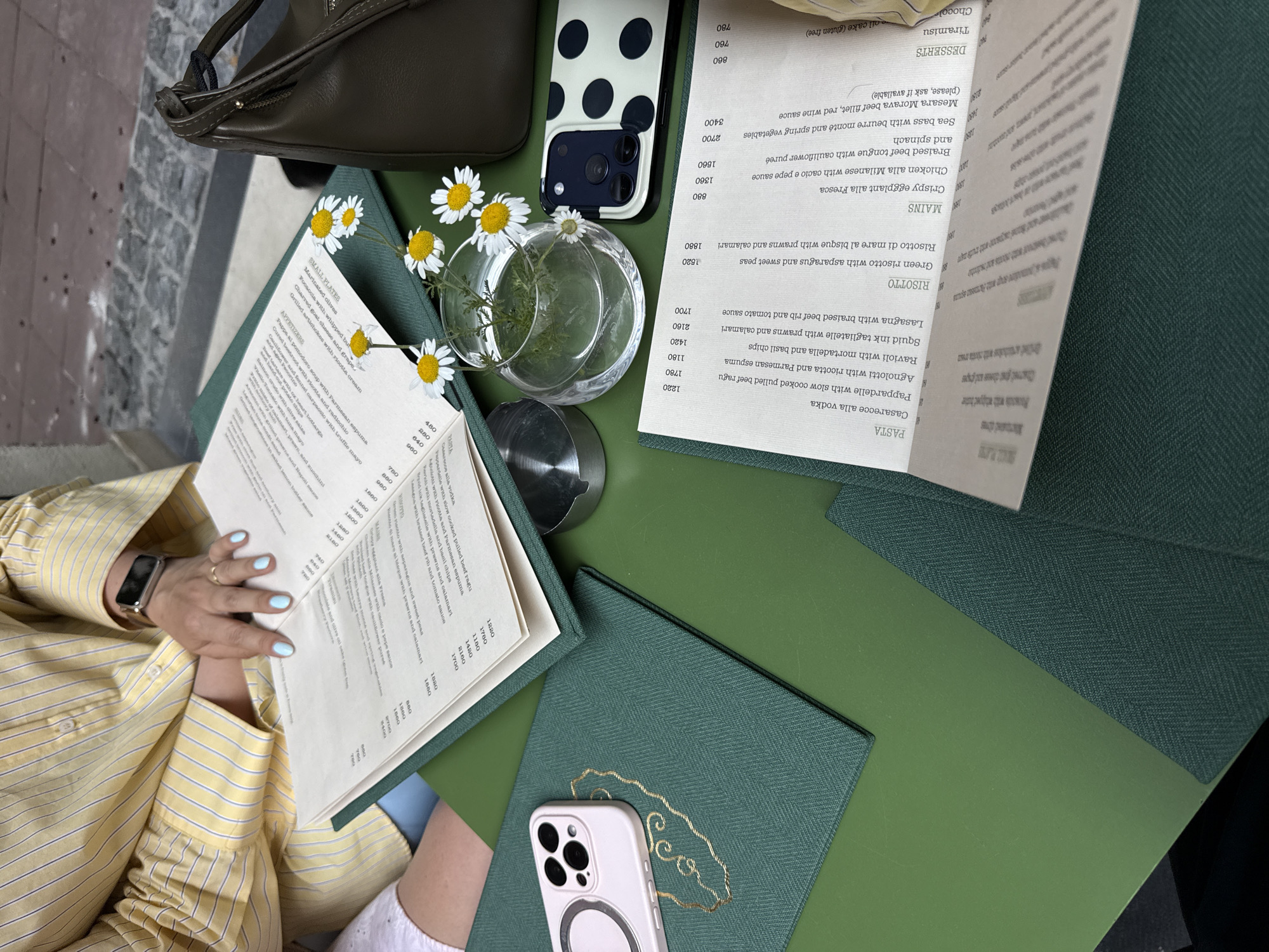 A high-angle, full shot depicts a restaurant table setting, likely in Belgrade, Serbia, given the "Leo" insignia on the menu, an establishment in that city. A person wearing a striped yellow shirt and an Apple Watch sits partially in the frame, their hands with light blue nail polish resting near the menu.
The table is covered with green fabric and holds several items. A white iPhone in a clear case with a magnetic ring lies near a closed menu. A second iPhone, in a white case with black polka dots, sits next to an olive-green handbag. A small, round glass vase contains several white daisies. Open menus reveal Italian dishes, including risotto and pasta, with prices in Serbian dinars. Silverware is partially visible near the vase. The table appears to be outdoors on a light-colored surface, possibly pavement.