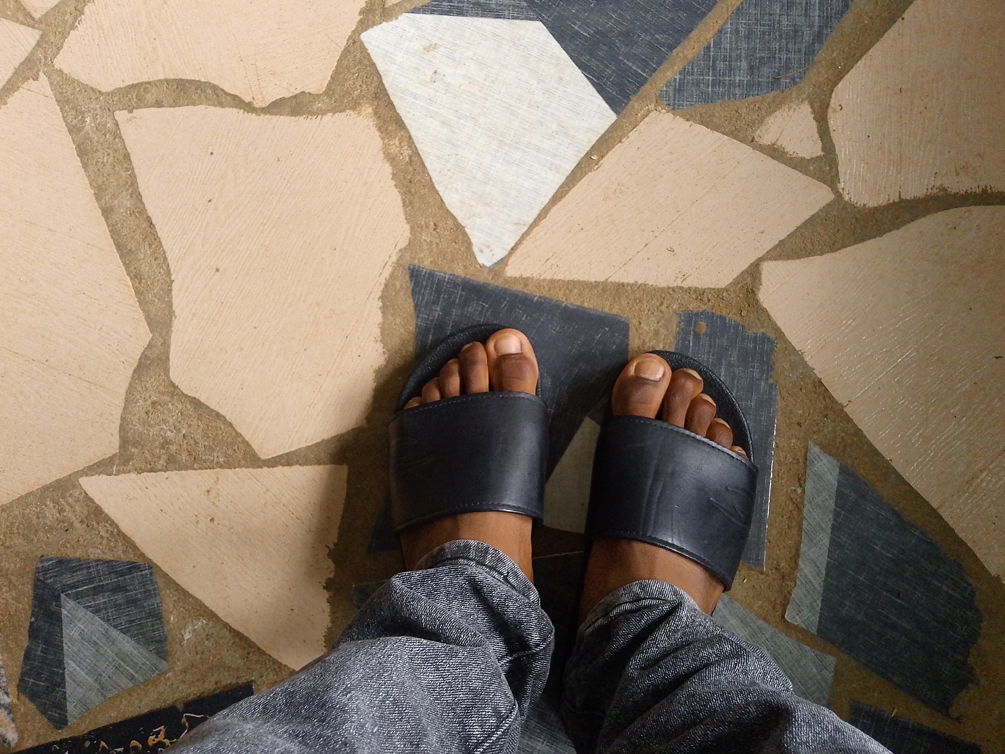 The image shows a top-down view of a person's feet wearing black slide sandals and denim jeans. The feet are positioned on a floor tiled with irregular-shaped pieces of varying colors, primarily light beige, white, and dark gray with a textured pattern. The tiles are bordered by what appears to be grout or cement. The lighting suggests it is daytime, and the environment seems to be indoors or a sheltered outdoor area. There is no visible text in the image. The overall impression is casual, with the focus on the footwear and the unique flooring.