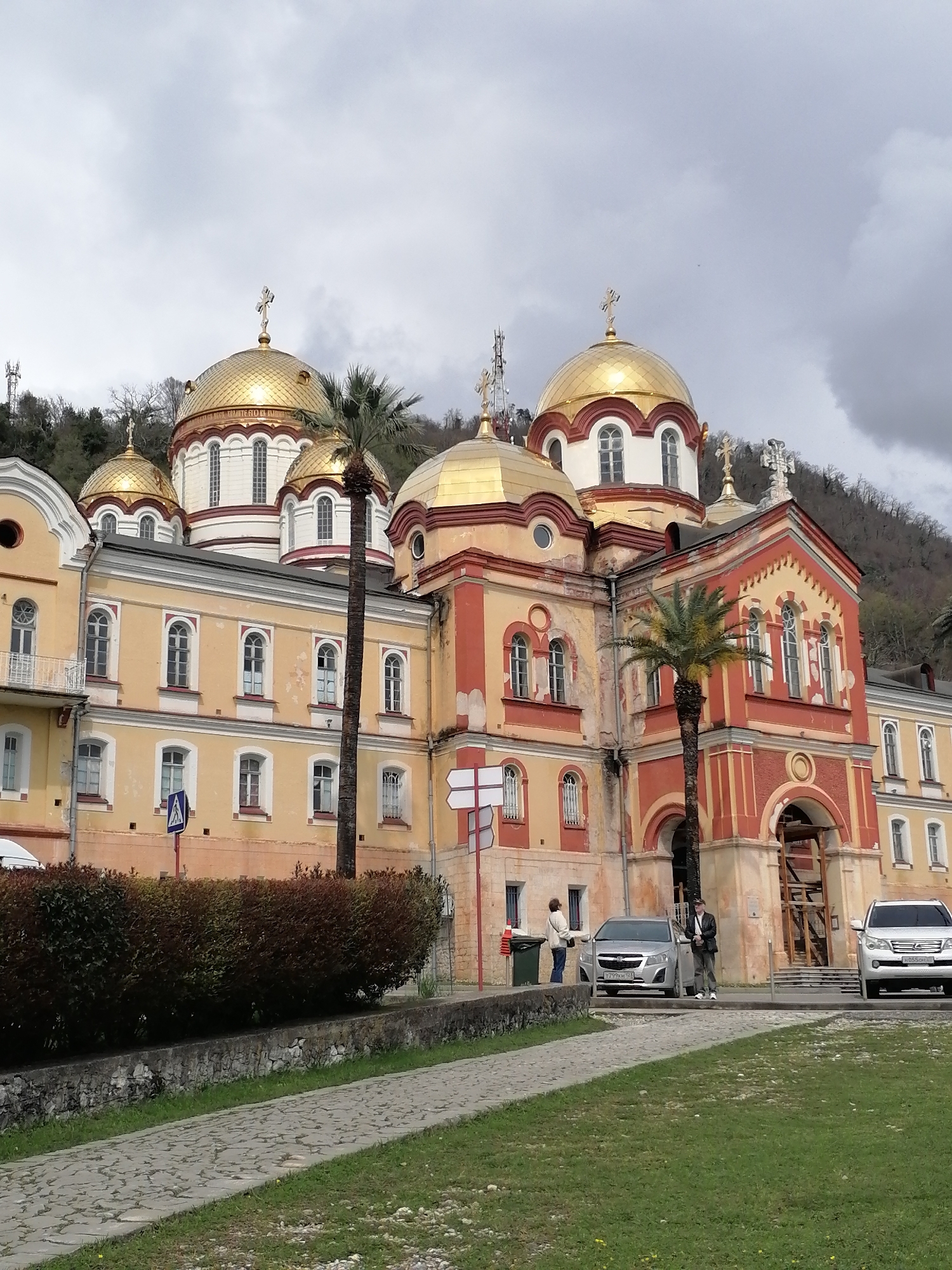 The image captures a grand Orthodox monastery or church, characterized by its multiple golden, onion-shaped domes topped with crosses, set against an overcast sky and a backdrop of a forested mountain. The building itself is multi-storied, primarily painted in shades of yellow and cream, with distinct red and orange decorative accents around windows and architectural details. Several palm trees are visible, suggesting a warm climate.
In the foreground, a paved path with a cobblestone-like texture leads to a grassy area. On the right side of the frame, two individuals are present near parked cars. One person, dressed in light clothing, stands by a green trash bin, while another, wearing a dark jacket and cap, stands beside a silver car. A white SUV is parked further right. Faint Cyrillic text, appearing to be "ТВОР" or part of a similar word, is visible on one of the golden domes. A blue pedestrian crossing sign is also visible near a bush. The silver car displays a Russian license plate "T799KH 102," and the white SUV has a partial Russian plate "H055OO 1_".
The scene depicts a daytime setting under cloudy skies, with no direct sunlight. Construction or maintenance scaffolding made of wood is noticeable near an arched entrance on the right side of the main building. The presence of palm trees, the architectural style, and the Russian license plates strongly suggest a location within Russia or a region with strong Russian cultural ties, such as Abkhazia.