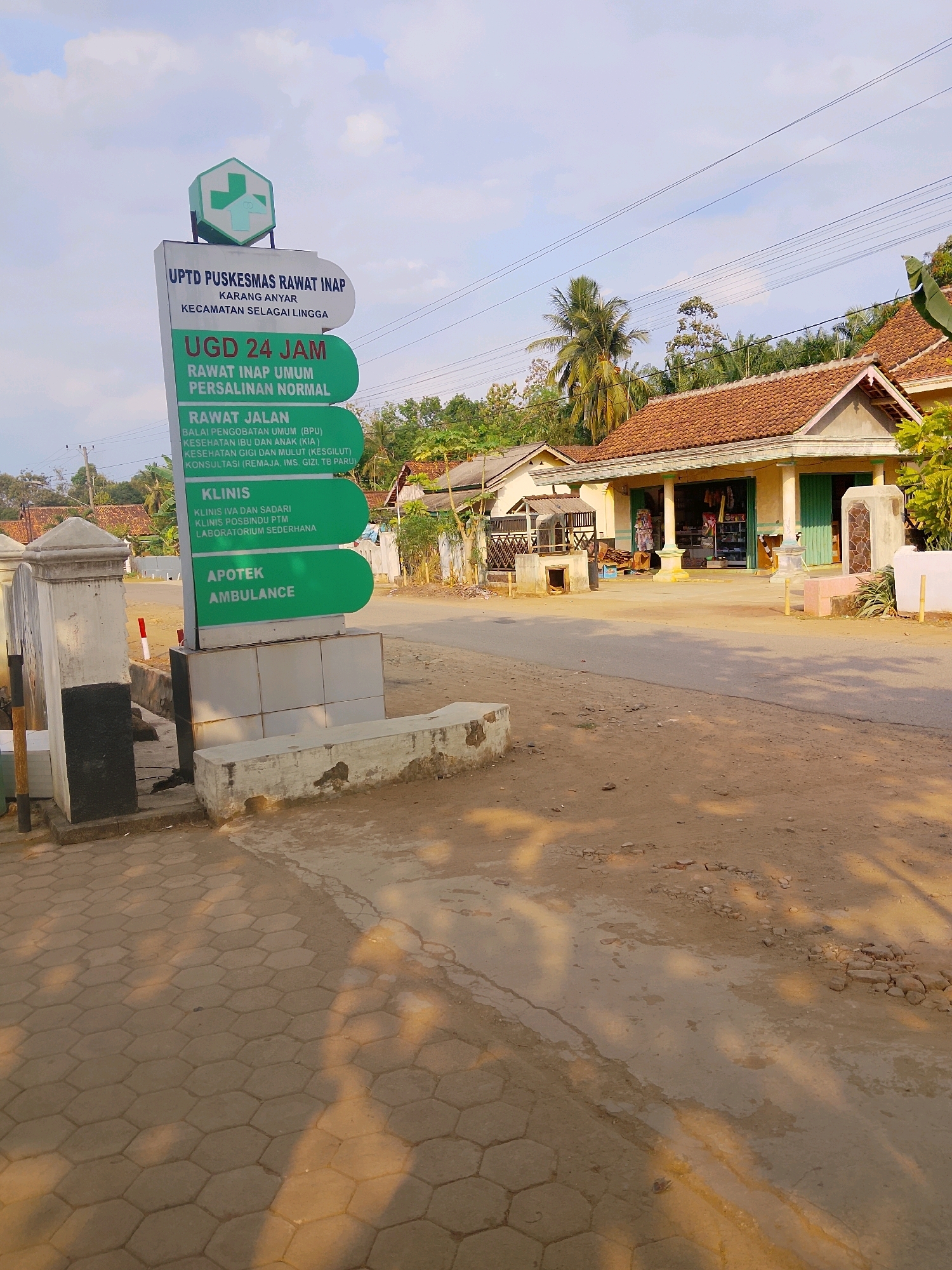 The image depicts a street scene in Negeri Agung, Indonesia, in front of a UPTD Puskesmas Rawat Inap (Inpatient Community Health Center). A tall, multi-sectioned green sign on the left side of the frame provides information about the facility's services. The top section of the sign features the Puskesmas logo, a white cross within a green hexagonal outline. Below this, text indicates "UPTD PUSKESMAS RAWAT INAP", "KARANG ANYAR", and "KECAMATAN SELAI LINGGA". Further down, sections are dedicated to "UGD 24 JAM" (24-hour Emergency Room), "RAWAT INAP UMUM" (General Inpatient Care), "PERSALINAN NORMAL" (Normal Delivery), "RAWAT JALAN" (Outpatient Care) with sub-categories like general medical consultation, maternal and child health, dental and oral health, and consultations for adolescents, chronic diseases, and tuberculosis. The bottom sections list "KLINIS" (Clinic) with specific types of clinics and "APOTEK AMBULANCE" (Pharmacy Ambulance).
To the right of the sign, a paved area with hexagonal tiles leads to a road. The road itself is dusty and appears to be a rural or semi-urban street. Beyond the road, there are several modest houses with terracotta tile roofs. One house in the mid-ground is open at the front, resembling a small shop, with goods displayed inside. Tall trees, including palm trees, are visible in the background, suggesting a tropical or subtropical climate. The sky is partly cloudy. The overall impression is of a functional but modest community health facility in a village setting. No people are visible in the image.