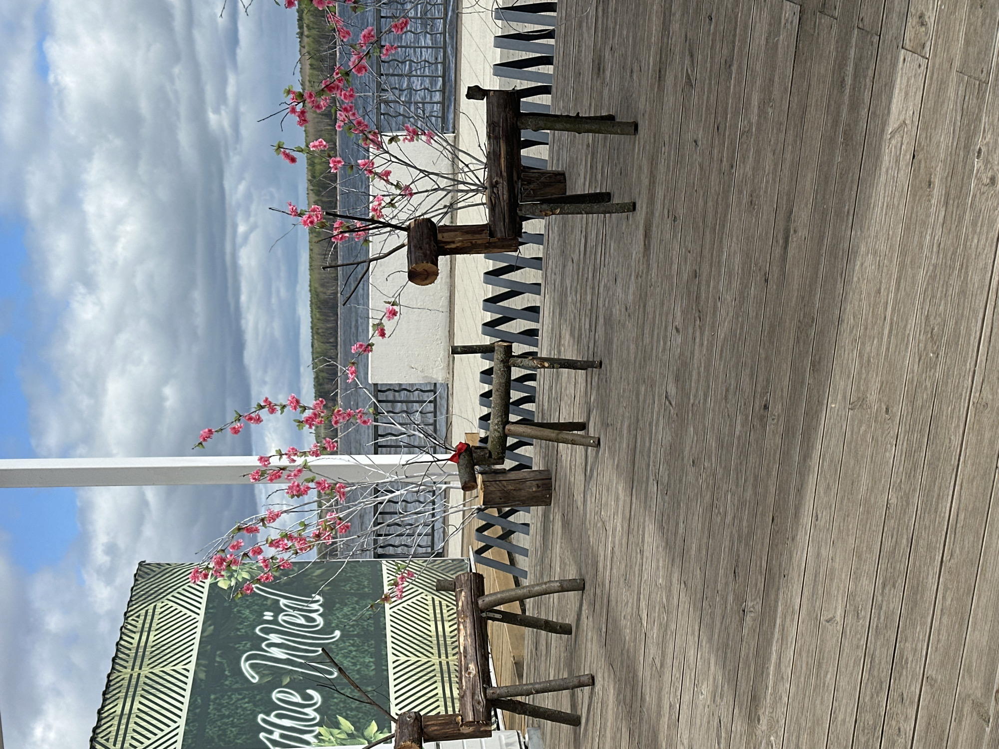 The image shows three wooden reindeer decorations with flowering branches placed on a wooden deck. The background includes a body of water, a railing, and a partly cloudy sky. To the left is a sign that reads "Cafe The Mid." The photo was taken in the city of Chaikovsky, Russia.