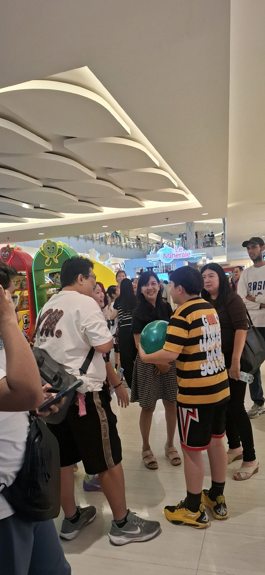 The image depicts a scene in a brightly lit indoor shopping mall, possibly in Grogol, Indonesia, with a focus on a group of people gathered around a display or promotional area. In the foreground, several individuals are visible, including a man in a white t-shirt and black shorts, a woman in a black dress, and a young person in a yellow and black striped shirt holding a green balloon. The ceiling features a modern, curved architectural design with integrated lighting. Behind the main group, other shoppers and mall patrons can be seen walking on the upper level, suggesting a bustling environment. Various colorful displays and signage are present in the background, hinting at a retail or promotional event. The overall impression is one of a social gathering or event taking place within a commercial space.