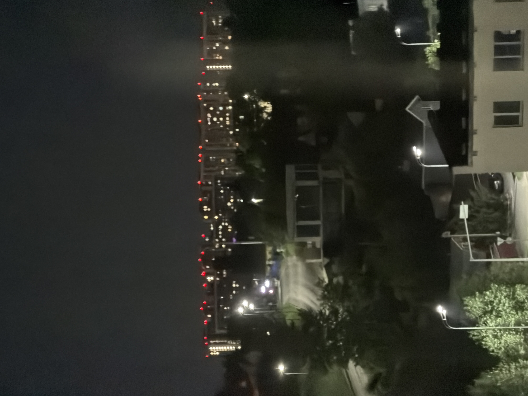 The image depicts a nighttime cityscape in Kazan, Russia, viewed from an elevated position. The scene is dominated by the dark sky and the illuminated windows of several buildings, suggesting a residential or office area.
In the foreground, a tall, slender building is visible, adorned with a string of red lights along its edge. This building's facade has multiple windows, many of which are lit, indicating occupancy. To the right, another building with a lighter facade features several rectangular windows.
The middle ground reveals a network of streets and pathways illuminated by streetlights. Trees are scattered throughout the scene, their foliage dimly lit by the artificial light. Some structures appear to be part of the urban infrastructure, possibly multi-story buildings or complexes.
In the distance, a line of taller buildings forms a skyline, their lights creating a sparkling effect against the dark sky. The overall impression is one of an active urban environment at night. The time of day is clearly night due to the darkness and artificial illumination. There are no people or specific events readily identifiable in the image. The weather appears clear. No visible text is present in the image.