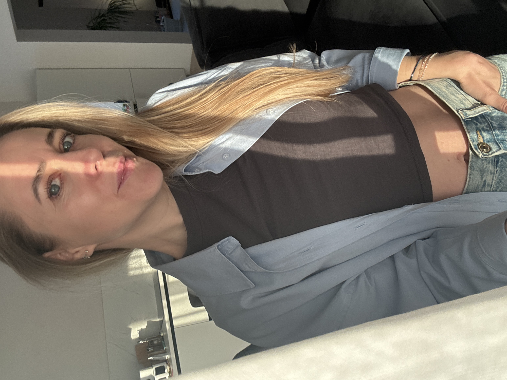 The media file displays a portrait-style photograph of a woman lying down or leaning, with her head positioned towards the left side of the frame and her body extending to the right. She has long, straight blonde hair that falls over her shoulders, and striking blue or green eyes that gaze directly at the camera. Her complexion is fair, and she has a calm, slightly smiling expression.
She is dressed in a dark brown or black crop top, exposing a section of her midriff. Over this, she wears an unbuttoned, long-sleeved light blue shirt, possibly linen, which is open and draped around her. Her lower body is clad in light-wash blue jeans. On her left wrist, she wears multiple thin, beaded bracelets, and a small silver stud earring is visible in her left ear.
The scene is an indoor setting, characterized by strong natural light entering from the upper left, creating bright highlights on her hair and face, and casting shadows across her clothing and the background. The background is softly out of focus, showing dark furniture, possibly a sofa, behind her head and upper body. Further back, light-colored walls or architectural elements are visible, along with a blurred hint of a green houseplant in the upper portion of the frame. The overall atmosphere is relaxed and casual, likely a daytime shot due to the bright sun. There is no visible text in the image.