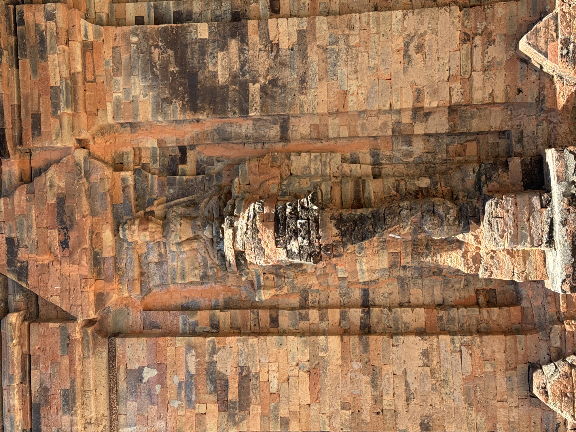 A full-shot, eye-level, exterior, medium-detail, vertical, medium-lighting, still image captures a brick temple in Nha Trang, Vietnam. The temple is constructed of reddish-brown and dark grey bricks, arranged in a horizontal pattern. The bricks are weathered and show signs of age, with some discoloration and chipping. In the center of the image, a carved figure of a deity is embedded in the temple wall. The deity is depicted seated with legs crossed and hands resting on its lap. It wears an elaborate headdress and is adorned with jewelry. The deity's face is serene and contemplative. The surrounding brickwork is textured and appears to be aged and weathered. The sunlight casts shadows on the temple, highlighting the intricate details of the carvings and the texture of the bricks. The overall impression is one of ancient grandeur and spiritual significance.