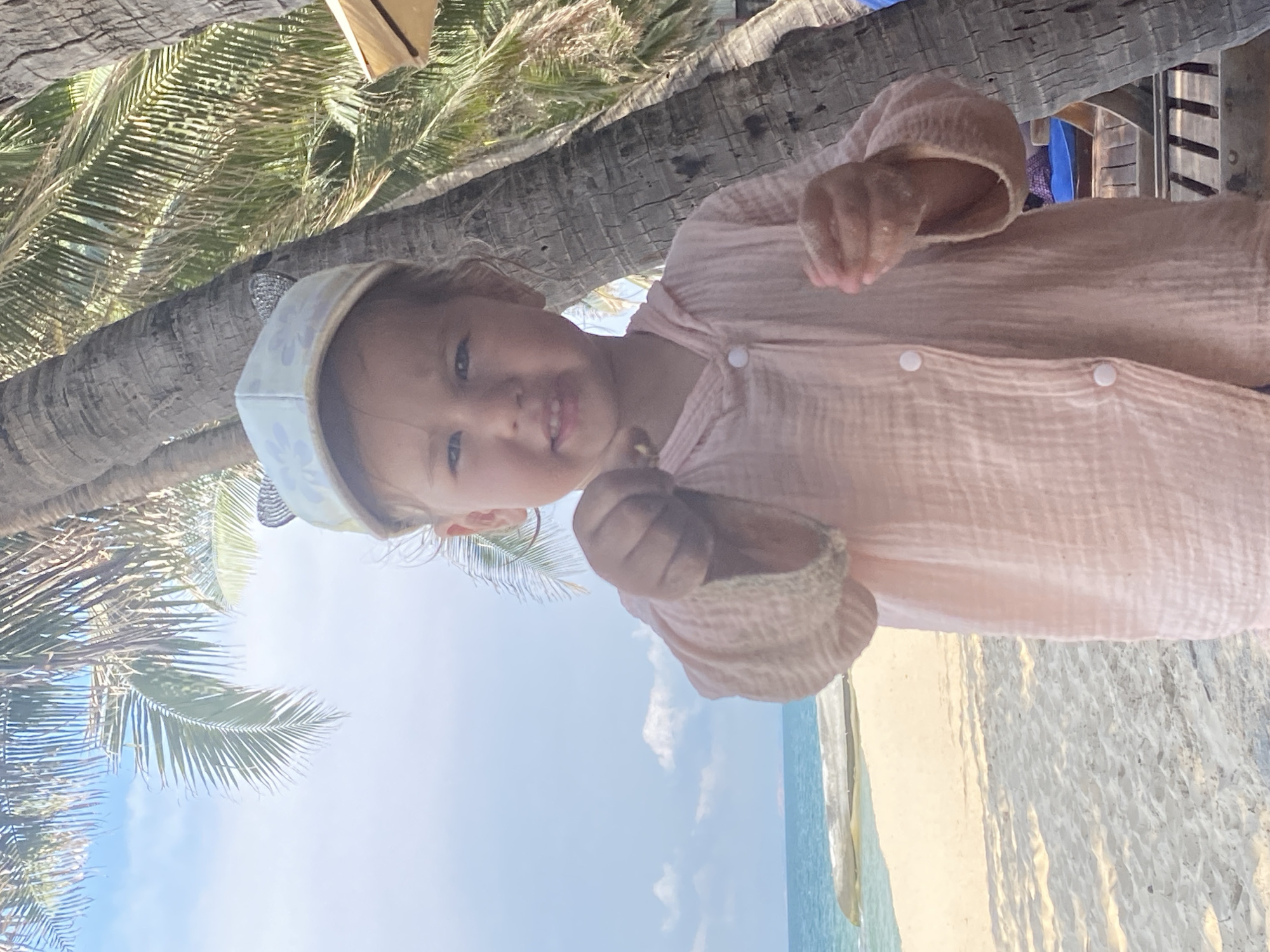 A young girl, wearing a pale pink hooded button-up shirt and a light blue cap with floral patterns and cat ears, stands on a sandy beach in front of palm trees. She is holding a small object in her right hand, which is extended towards the camera. The beach has a strip of sand leading to the turquoise ocean, with some clouds in the bright blue sky. Behind her, to the right, is the trunk of a large palm tree, and to the left, more palm fronds. The scene appears to be in Phu Thuy Ward, Vietnam.