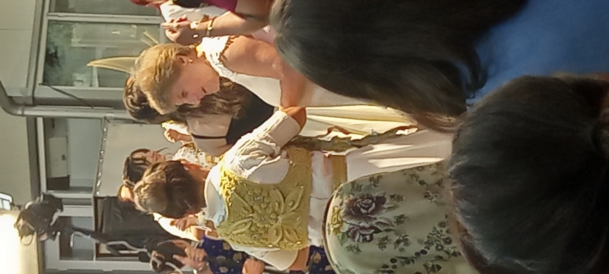 In Lyuben, Bulgaria, a group of people are gathered in what appears to be an outdoor or semi-outdoor event. The foreground is dominated by the backs of several individuals, two of whom are wearing patterned headscarves. The middle ground features a woman in a white dress with a lace collar, holding hands with another person in a traditional-looking outfit consisting of a white shirt and an ornate, embroidered golden vest. Behind them, other individuals are visible, some in traditional attire and others in more casual clothing, with a backdrop of building structures and what looks like a camera on a stand.