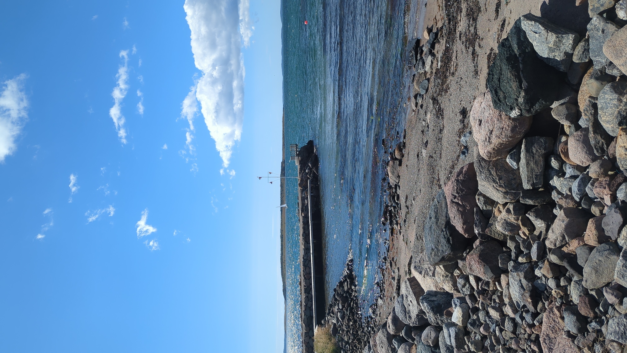 A vertical, eye-level, wide shot shows a rocky shoreline leading to a pier on the left and extending into a body of water under a blue sky with scattered white clouds. The foreground is dominated by an array of large and small rocks, with some sand and dried seaweed visible between them. The rocks are varied in color, including gray, brown, and pinkish hues. The water is a deep blue-green, with small waves creating ripples and reflecting sunlight. A wooden pier with a stone base juts out into the water from the left side of the frame. At the end of the pier, there is a small, open-air structure with a railing and a flagpole with two small flags. A single sailboat is visible in the distance to the left of the pier. To the right of the pier, a bright orange buoy floats on the water. In the far distance, a landmass with trees is visible on the horizon. The scene appears to be a coastal area in Larkollen, Norway, likely a public access point or small harbor.