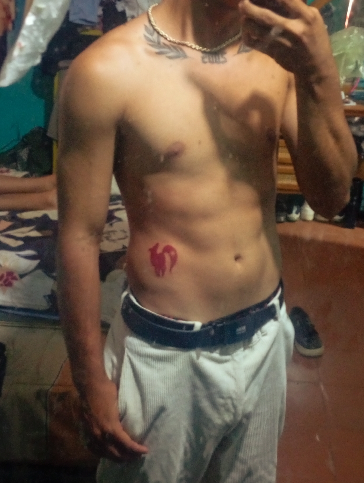 A shirtless man is standing indoors, taking a selfie. He is wearing white pants with a dark belt. He has tattoos: one of a symbol resembling a pair of foxes or wolves on his left side and another, partially obscured, on his chest. He is also wearing a gold chain. The location is unknown. The description doesn't specify an Altagracia, Venezuela location.