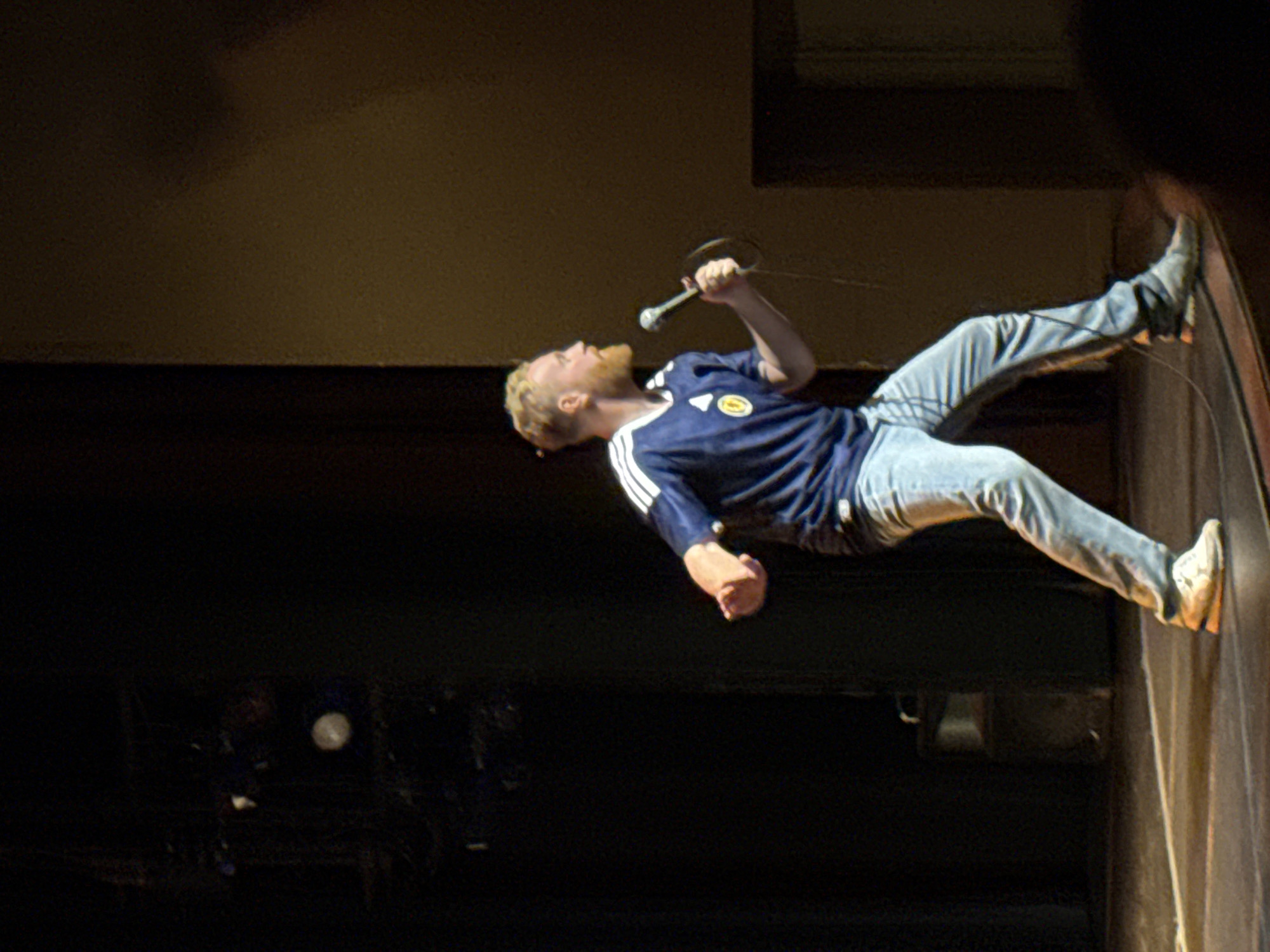 A fair-skinned male with blondish-red hair and a beard stands on a stage, holding a microphone in his right hand and speaking into it. He is wearing a blue t-shirt with white stripes on the sleeves and a circular emblem on the chest, and light blue jeans. He is also wearing white sneakers. The man is positioned in the center of the frame, facing left, with his body angled towards the audience. The stage is dimly lit, with dark curtains behind the performer and a spotlight illuminating him from the side. The backdrop appears to be a theater or performance venue. There are no other people or discernible objects in the immediate vicinity of the performer. The lighting and stage setup suggest a live performance.