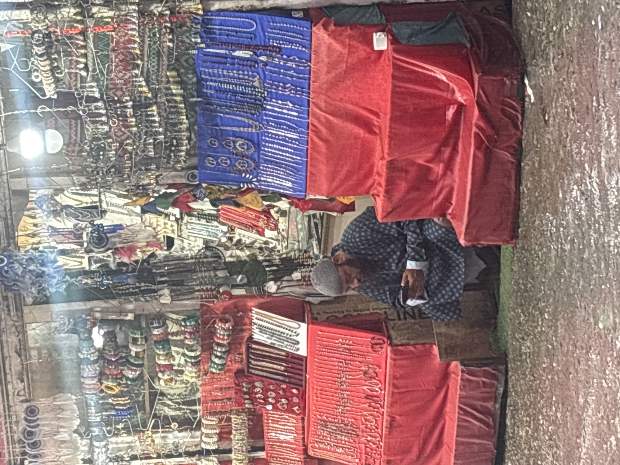 In Vashisht, India, a man wearing a grey cap and a patterned blue garment sits on the wet ground, looking at his mobile phone. He is positioned in front of a stall displaying a variety of jewelry. The stall features multiple levels of red fabric draped over stands, showcasing rows of necklaces and bracelets. Above this, two blue fabric panels are adorned with strings of beads and pendants. The background is filled with more stalls displaying an abundance of jewelry, trinkets, and other items hanging from racks and displayed on shelves. The ground is wet, indicating recent rain, and small puddles reflect the overhead lights of the market.