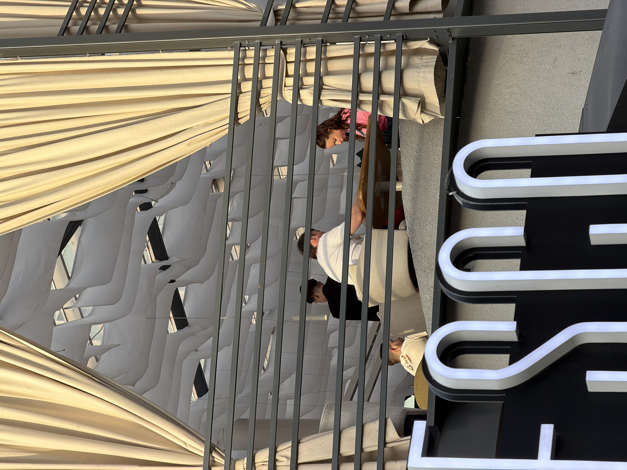 The image captures an outdoor or semi-outdoor dining area, likely a restaurant or cafe, in Krasnoyarsk, Russia, during daylight hours. The scene is viewed through a grey metal framework, possibly part of a pergola or balcony railing, which is draped with light beige fabric creating a ceiling and side coverings.
Several people are present: a woman in a pink top is seated at a table on the left, looking downwards. In the middle, a man in a white shirt and apron, likely a server or chef, stands looking towards the right. Another person with dark hair is visible behind him. Further right, a man in a white baseball cap is seated.
Beyond the immediate dining area, an artistic ceiling design features undulating white fabric or sculpted panels with dark, rectangular cutouts, giving the space a modern aesthetic. Bright, natural light illuminates the scene, casting soft shadows and highlighting the texture of the drapes.
On the far right, a large sign with illuminated white lettering against a black background is partially visible. The discernible shapes form parts of letters, suggesting "THE". The setting appears relaxed and bright.