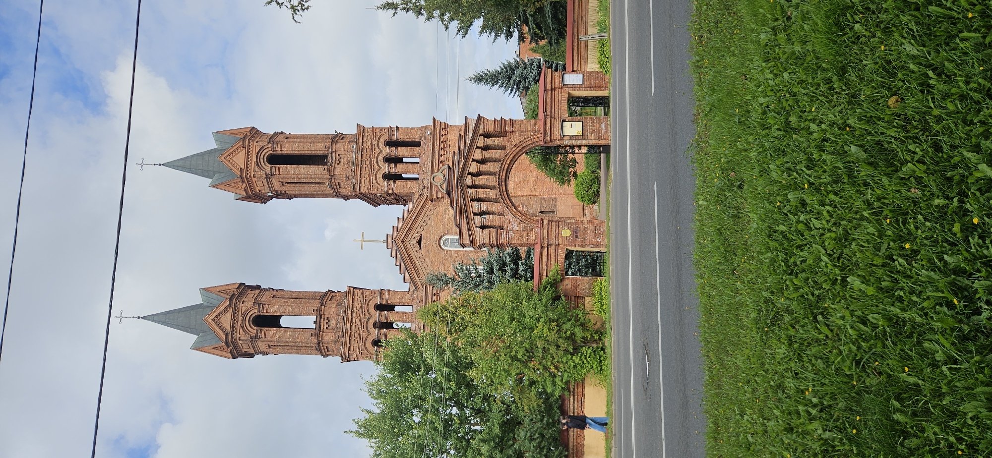 The image captures a two-towered brick church with a central arched entrance, located in Vitsebsk, Belarus. The church features Gothic architectural elements, including pointed spires topped with crosses. The facade is constructed from red brick, with decorative brickwork detailing. Green trees and a low brick wall with an iron gate surround the church grounds, separating it from a paved road in the foreground. A person in dark clothing and blue jeans is walking along the sidewalk next to the wall, moving from left to right. The sky is partly cloudy, with blue patches visible. The foreground is dominated by a well-maintained green lawn.