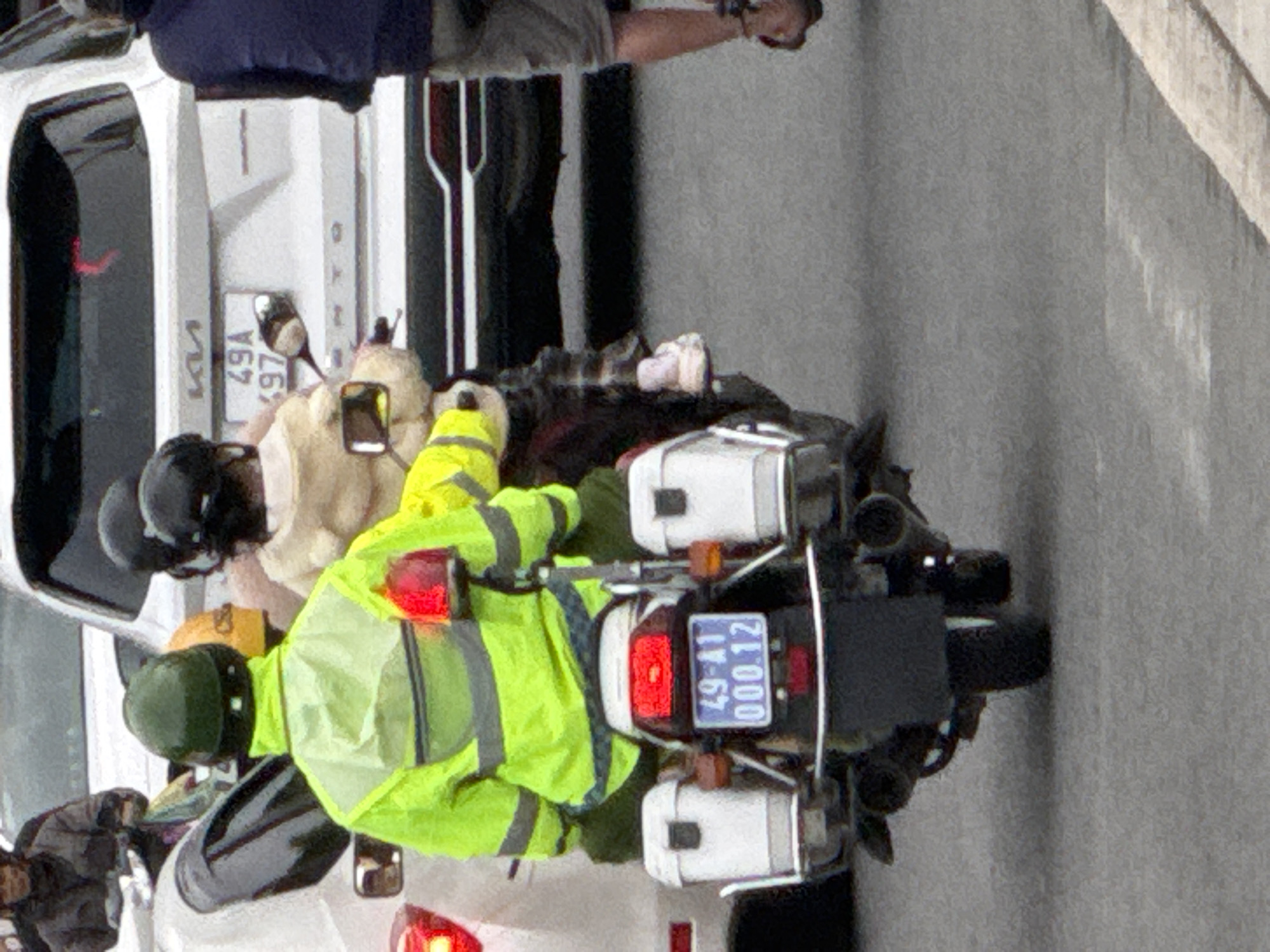 The image depicts a street scene in Da Lat, Vietnam, with a focus on a motorcycle and its riders. In the foreground, a person wearing a bright yellow reflective jacket and a green helmet is seated on a motorcycle. The motorcycle has two white storage boxes mounted on either side of the rear, and a license plate that reads "49-A1 000.12". Behind this motorcycle, another motorcycle is partially visible with a person in a tan jacket. Further in the background, a white Kia Sportage SUV is parked on the street. The overall environment suggests an urban setting, likely a street in Da Lat, characterized by the presence of vehicles and pavement. The image captures a moment of urban traffic.