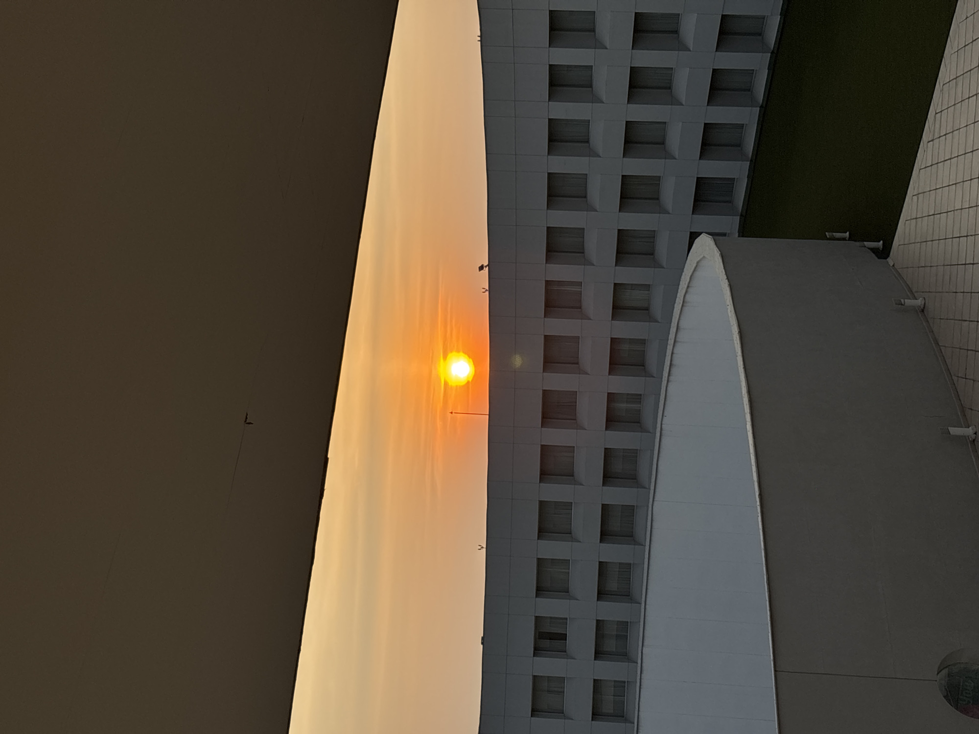 Captured from a partially obscured vantage point in Mexico City, Mexico, the image showcases a vibrant sunset framed by architectural elements. To the left, a blurred, dark expanse, possibly a building's edge, contrasts sharply with the bright orange and yellow hues of the setting sun. The sun, positioned centrally, illuminates a smoky, hazy sky. To the right, a multi-story building features a grid-like pattern of windows, juxtaposed against a rounded, concrete structure. A patch of green is visible, suggesting a small lawn or garden area. The orientation of the image is rotated, requiring the viewer to adjust their perspective.