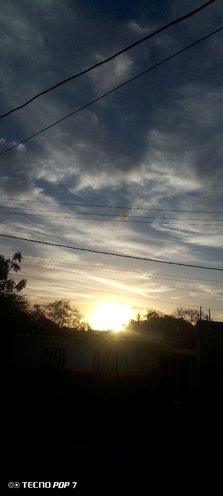 The image shows a sunset in Altagracia, Venezuela. The sky is filled with dark and light clouds and a few wires cut across the sky. The sun is obscured by trees and buildings, creating a silhouette effect. The foreground is dark, suggesting the image was taken in low light conditions. The bottom of the image includes "TECNO POP 7".
