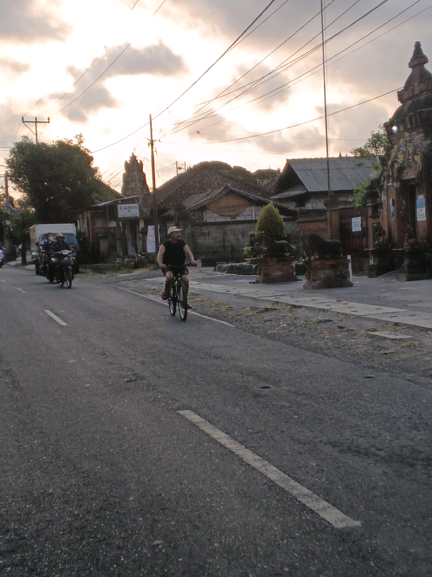 In Sukawati, Indonesia, a man wearing a tan hat and a dark t-shirt rides a bicycle down a paved street. He is in the foreground, to the right of the frame, with his body angled away from the camera. Behind him, two motorcycles are approaching from the distance, one with two passengers wearing masks and the other with a single rider. The motorcycles are on the same paved road, with the cyclists in the foreground. The road is lined with buildings and structures, some of which appear to be shops or temples, indicating an urban or semi-urban environment. A large white truck is parked on the left side of the road. The sky is overcast with dramatic clouds, suggesting either sunrise or sunset. The overall scene depicts a street view in Sukawati during twilight hours, with ordinary daily activities like cycling and motorcycling taking place.