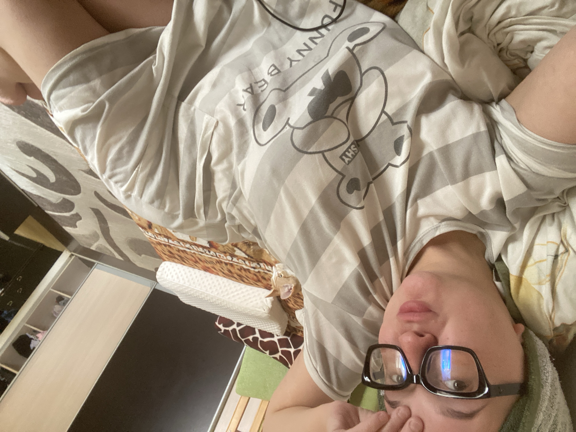 A young woman with light skin and short brown hair, wearing glasses and a striped t-shirt with a cartoon bear print, lies on a bed in a bedroom. The t-shirt has the text "FUNNY BEAR SHY" printed on it. The woman's right hand is touching her forehead. The room has a wardrobe with open shelves and various items on display. The bed has patterned bedding and a white textured pillow. The scene appears to be indoors, likely in a residential setting. The lighting suggests it could be daytime. There are no obvious indications of the weather. The overall mood appears relaxed and casual. The location is identified as Chaikovsky, Russia.