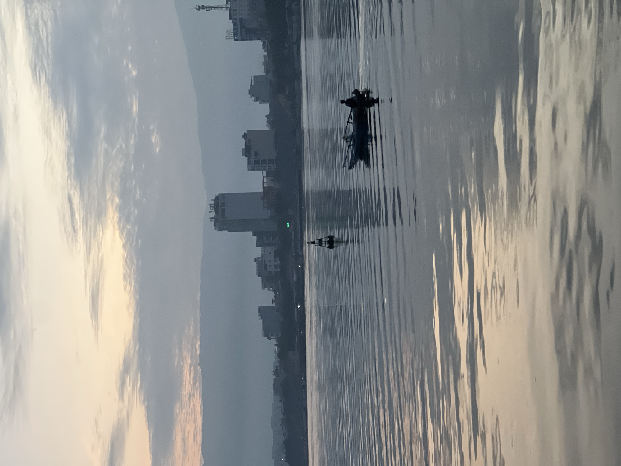 A wide shot of the Han River in Da Nang, Vietnam, shows the city skyline shrouded in a light mist under a cloudy sky at dawn or dusk. A lone person is visible in a small boat in the foreground, navigating the calm waters. A navigation buoy stands further out in the river. The water reflects the soft, muted colors of the sky and the buildings.