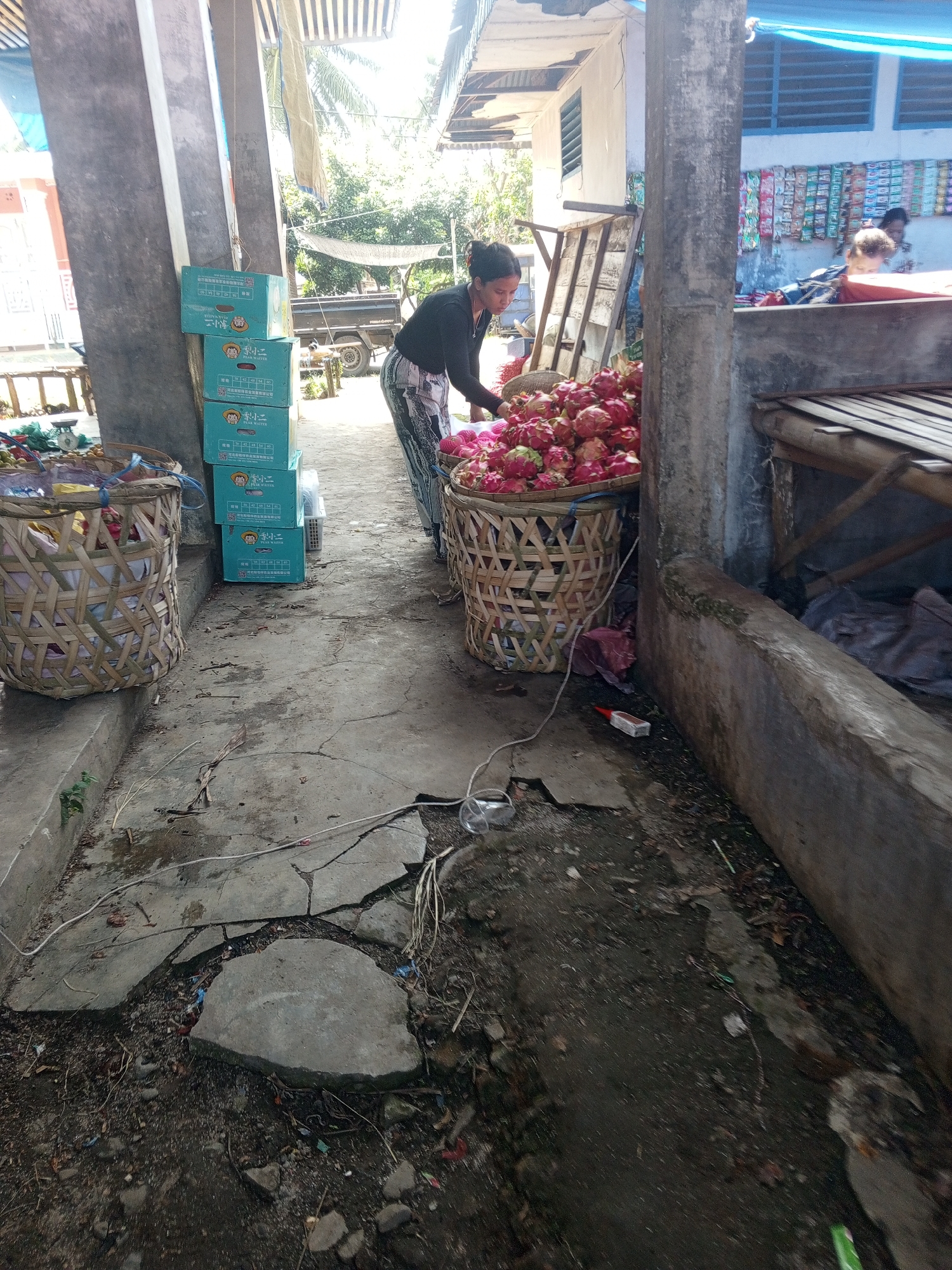 A woman is standing at a market stall in Talun Kenas, Indonesia, sorting through a large basket of dragon fruit. The stall is made of concrete pillars and a wooden roof structure. In the background, there are stacked boxes of goods and a display of packaged snacks. The ground is paved with concrete, which is cracked and uneven in places, with debris scattered around. To the right, a raised wooden platform suggests another market stall area. The scene appears to be an outdoor market, with natural light filtering through the roof.