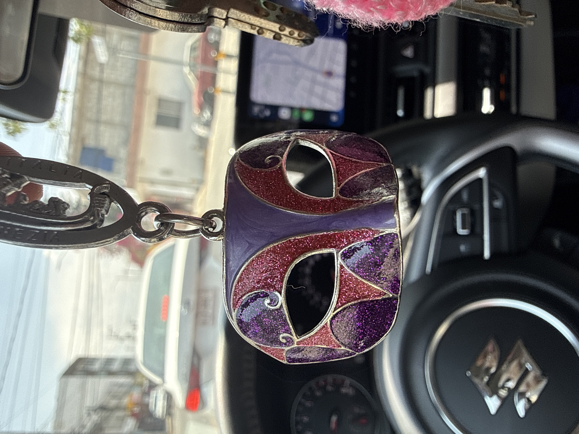 A close-up, eye-level shot captures a decorative keychain hanging inside a car in Puebla, Mexico. The keychain features an intricate, abstract design, possibly resembling a stylized letter or mask, filled with glittery purple and reddish-pink enamel, outlined in silver-toned metal. It is attached to a metal chain, which has the word "ITALIA" and partial text "REZIA" (likely part of an Italian place name) engraved on it.
The immediate background reveals the car's interior, including a portion of a black steering wheel with a prominent silver-colored Buick logo. Behind the steering wheel, an infotainment screen displays a map or navigation system. A small pink, fuzzy object is partially visible at the top edge of the frame.
Through the car's windows, blurred reflections of an outdoor scene are visible, indicating it is daytime with bright sunlight. The reflections show parts of buildings, suggesting an urban or suburban setting, and faint outlines of other vehicles, including a white van or SUV. The overall impression is a personal item displayed within a vehicle during daylight hours in Puebla, Mexico.