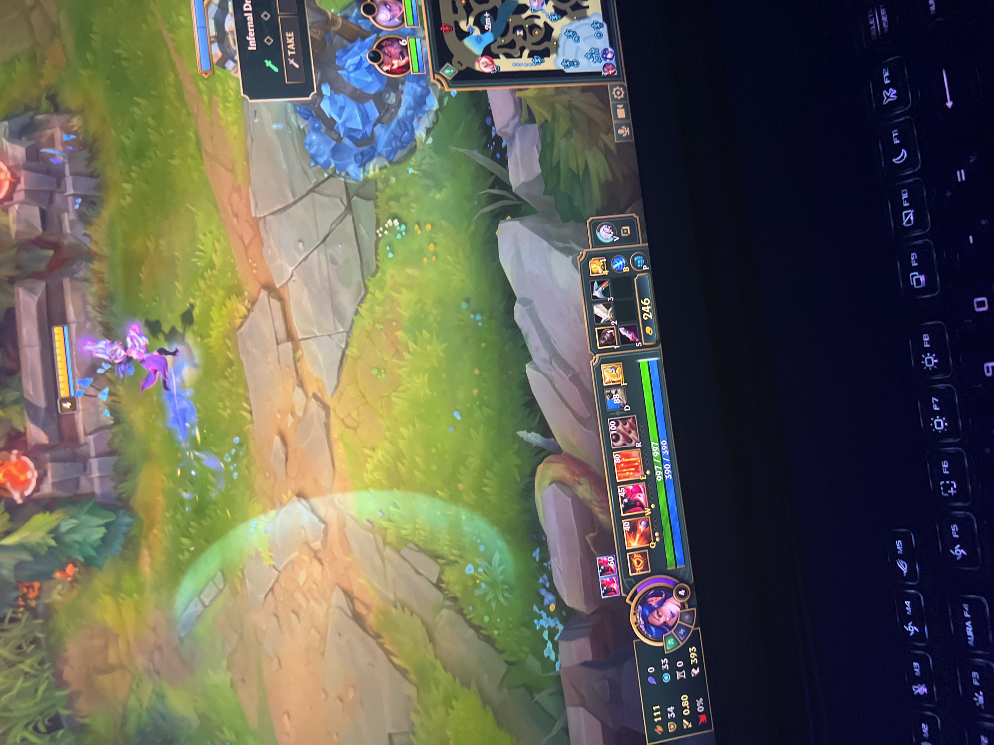 The image shows a top-down view of a video game, specifically League of Legends. The game takes place in a fantasy environment with grassy plains, rocky paths, and trees. In the center of the screen, a purple character with wings is casting a blue spell. On the right side of the screen, there is a mini-map showing the game's layout and player locations. The bottom of the image shows the game's user interface, including player statistics, abilities, and item slots. A portion of a gaming keyboard is visible at the bottom, with illuminated keys. The overall scene depicts an active game in progress.