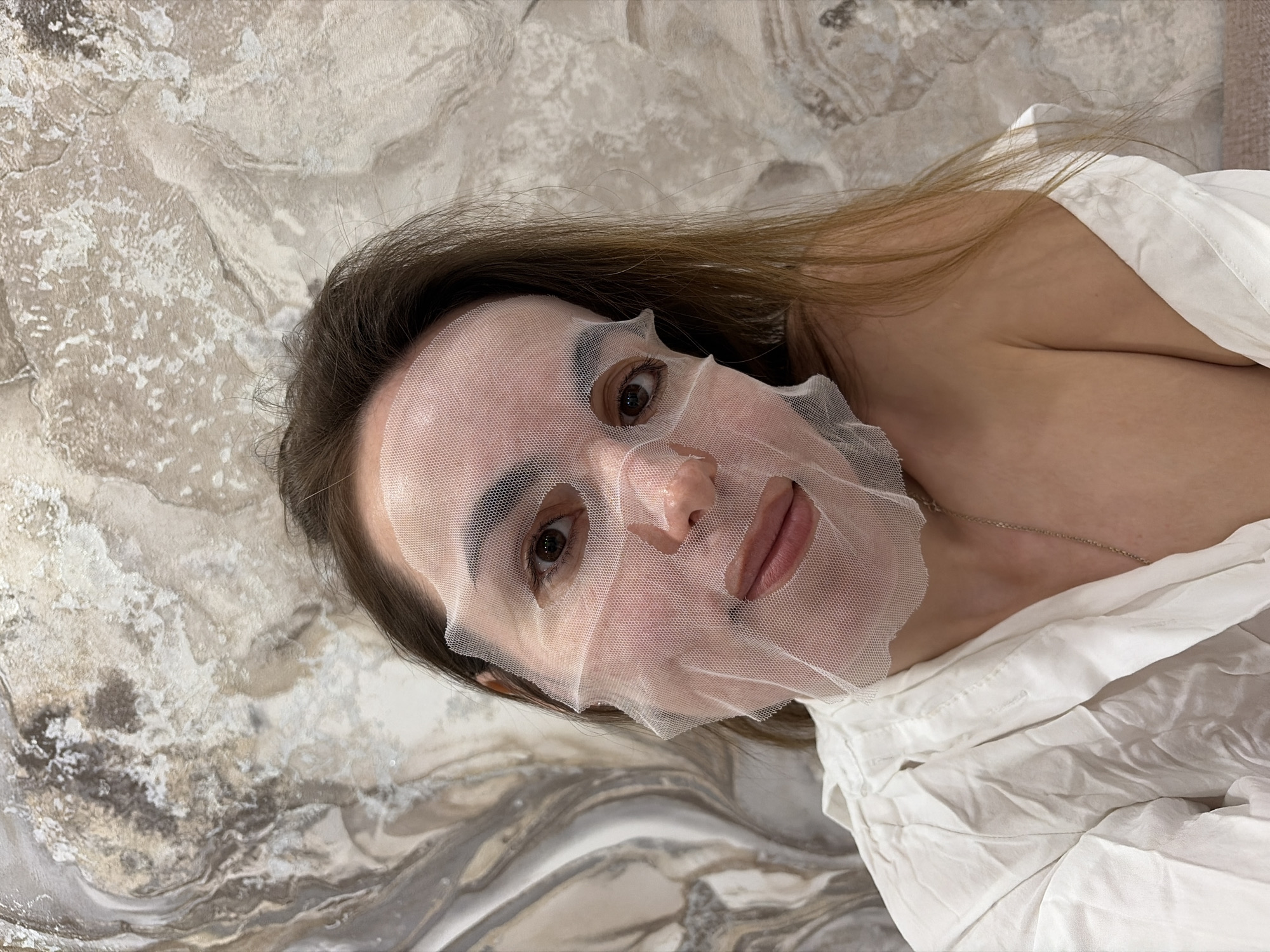 A woman is lying down, wearing a white shirt and a white facial sheet mask with holes cut out for her eyes. A silver chain necklace is visible around her neck. The background appears to be a textured surface resembling marble. The city of Izhevsk, Russia is not visible in the image.
