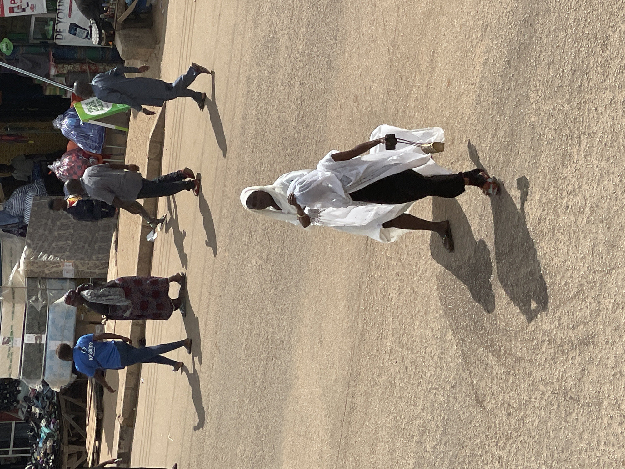 In a street scene, two women dressed in white Islamic attire walk on a dusty, paved road. The woman on the left wears a headscarf and a flowing white dress, while the woman on the right, partially obscured, wears a black garment beneath her white headscarf and outer robe, carrying a phone in her right hand. Their shadows are cast on the ground. In the background, several men are also walking, one in a blue t-shirt and jeans, another in a grey shirt and trousers, and a third in a dark blue traditional outfit. The setting appears to be a marketplace, with various goods stacked behind a low wall, including mattresses, bags, and what looks like shoes or sandals. A green sign that reads "VOICE ADATA PLANS" is visible. The time of day is sunny, indicated by the strong shadows.