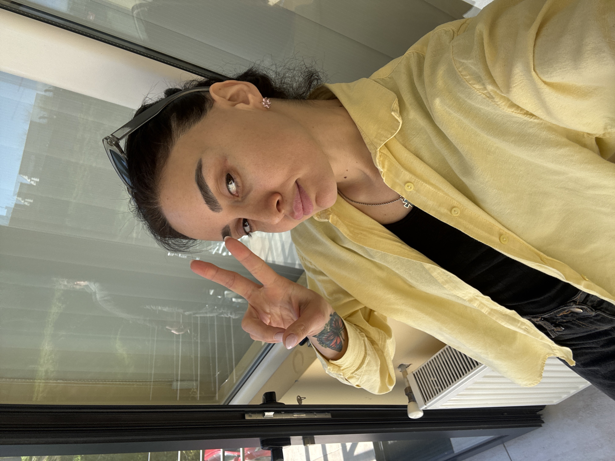 A young adult woman with dark hair pulled back, sunglasses perched on her head, and fair skin is posing indoors while making a "peace" sign with her right hand. She is wearing a yellow button-down shirt over a black top and dark jeans. Her left wrist has a butterfly tattoo. She has pierced ears with small flower earrings. She is standing by a window, reflecting a vague image of what appears to be city buildings. The window has a black frame and is possibly part of a balcony or enclosed porch. A white air conditioning unit or vent is beneath the window. Given the reflective buildings, the photo appears to be taken indoors, in a high-rise building, in Tbilisi, Georgia, as inferred from the query context.