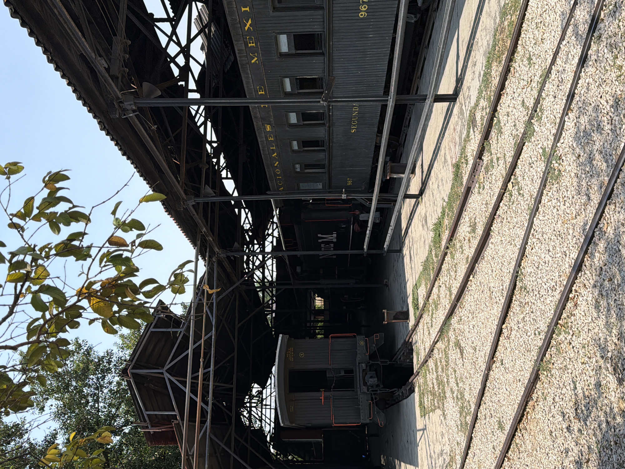 The image depicts a section of a railway depot in Cuautla, Mexico. The shot is taken from ground level looking upwards, showing railway tracks covered in light-colored gravel. Above the tracks are several old, dark-colored railcars under a decaying metal canopy structure. The railcars bear text that reads "NACIONALES DE MEXICO" and "SEGUNDA". The number "967" is visible on one of the railcars. Parts of trees with green leaves are visible on the left side of the frame.