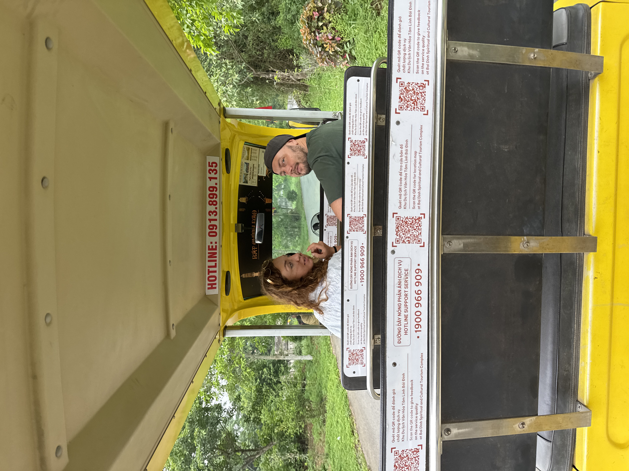 The image shows a yellow electric vehicle, likely a golf cart, in an outdoor setting with lush greenery and trees. Two people are seated inside the vehicle. A woman with curly brown hair is in the passenger seat, looking to her right. A man wearing a dark baseball cap and a dark green t-shirt is in the driver's seat, looking towards the camera. The vehicle has a white sign on the back with red lettering that reads "HOTLINE: 0913.899.135". Below the hotine number, there are multiple QR codes and Vietnamese text providing instructions to scan for location maps and service feedback. The text also mentions "Bai Dinh Spiritual and Cultural Tourism Complex," indicating the location. The overall scene suggests a tourist transport vehicle in a natural or park-like environment within the Bai Dinh Spiritual and Cultural Tourism Complex, which is located in Tây Hoa Lư Ward, Vietnam.