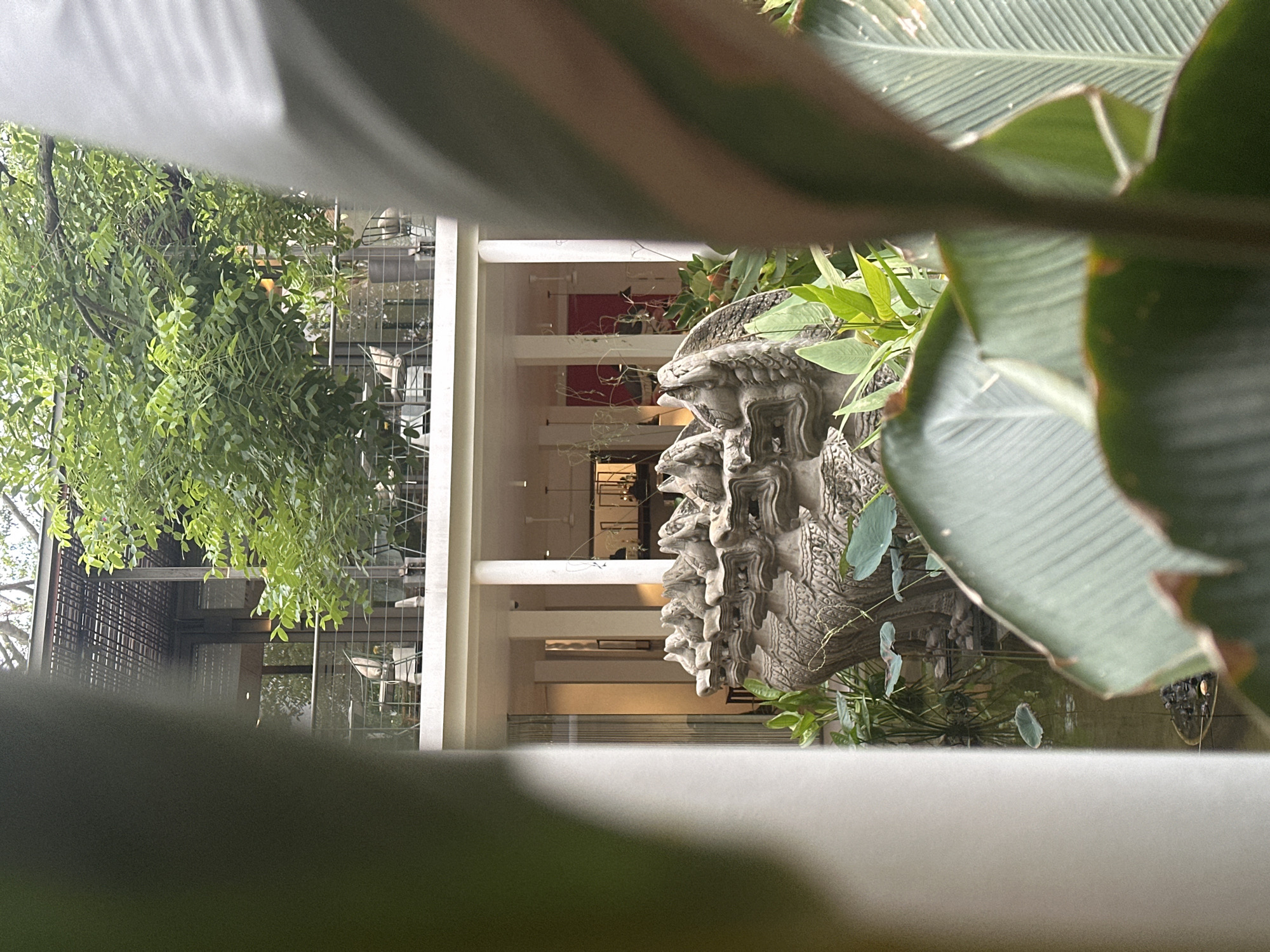 The image depicts an outdoor setting within the city of Khan Daun Penh, Cambodia. In the foreground, blurred large green leaves partially obscure the view. Behind these leaves, a stone sculpture of multiple mythical serpent heads is visible, situated near a pond with lily pads. In the background, a multi-story building with an open-air upper level featuring tables and chairs can be seen. The upper level is covered by a slatted roof structure. Lush green foliage surrounds the building. The scene appears to be a tranquil, possibly resort or restaurant, environment. There are no people clearly visible in the image.