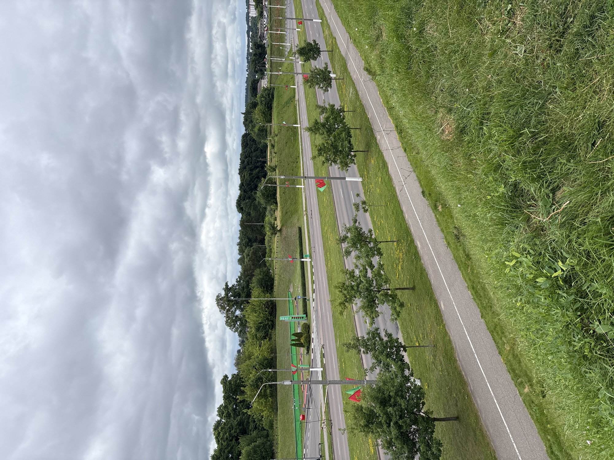 The image depicts an overcast day in Vitsebsk, Belarus, focusing on a multi-lane road and adjacent green spaces. In the foreground, a grassy embankment slopes upwards, with a paved pedestrian or bicycle path running along its base. A line of young trees with green foliage is planted between the path and the road. The road itself has multiple lanes marked with white lines, and a bike lane is visible on the far side. Red and green flags are mounted on poles lining the road at intervals. Beyond the road, a sloped, grassy hill rises, dotted with more trees. At the top of this hill, a bright green structure, possibly a playground or sports facility, is visible. In the distance, more trees and buildings, including some taller structures, are discernible under the cloudy sky. The overall impression is of a well-maintained urban or suburban area with a focus on greenery and infrastructure.