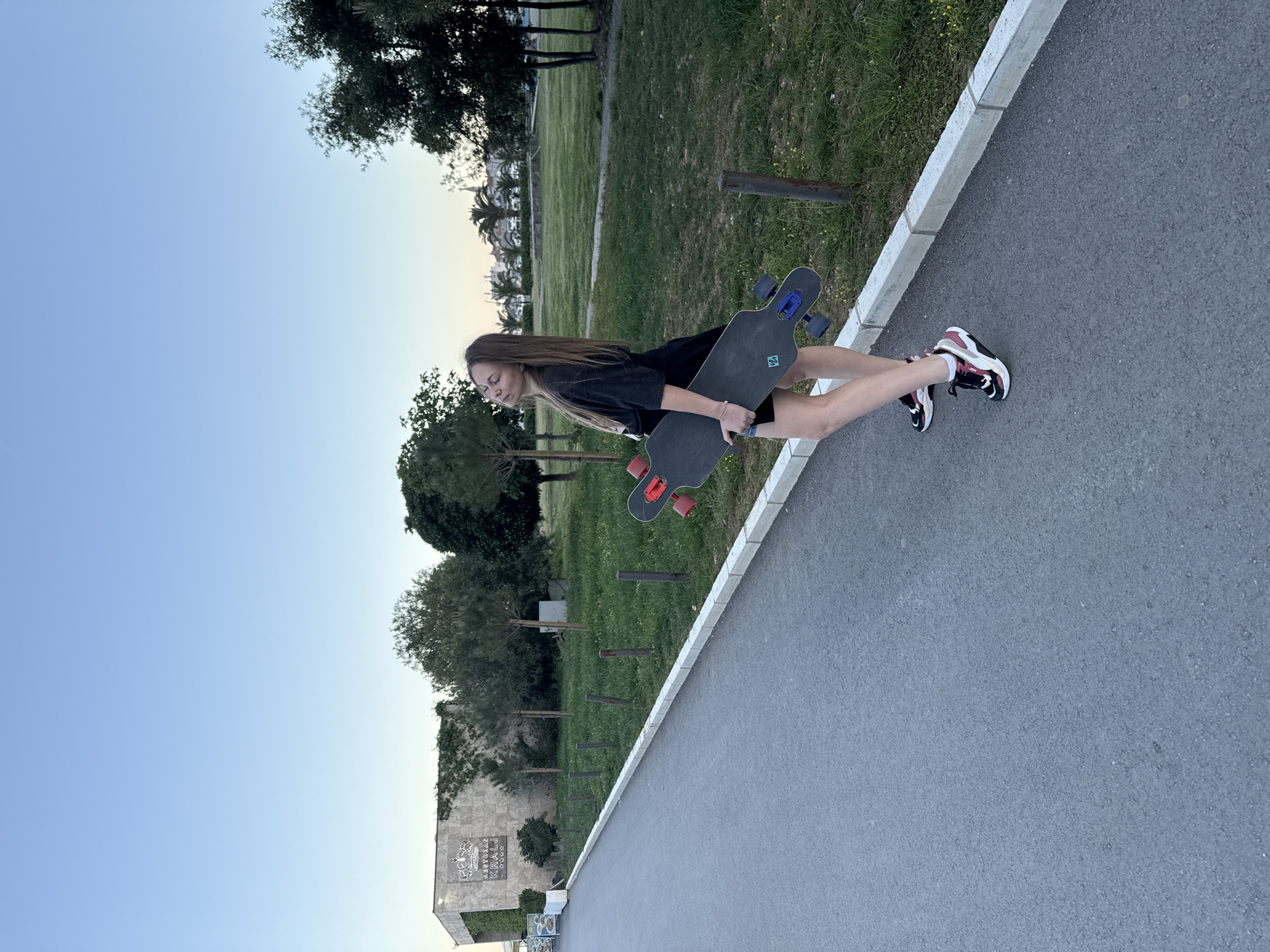 An outdoor, eye-level shot captures a young woman standing on a paved asphalt path, holding a longboard. The image is oriented such that the path runs diagonally from the bottom right to the middle left. To the left of the path, a concrete curb separates it from a grassy area dotted with trees and wooden posts. The woman, with long brown hair, wears a black t-shirt, dark shorts, and multi-colored sneakers (black, white, and red accents). She holds a dark longboard with blue trucks and red wheels.
In the midground, beyond the grassy patch, there are buildings and more trees. On the right side of the background, a large stone-faced building is visible, featuring a prominent crown logo and the text "RESTORAN KRALJ" with smaller text possibly reading "AZUR" below it. Further in the background, particularly on the left, additional buildings with red roofs and palm trees can be seen, hinting at a coastal town setting.
The lighting suggests late afternoon or early evening, with a clear, soft blue sky that transitions to a warmer, lighter hue near the horizon. The overall impression is calm and relaxed, with the woman appearing contemplative or resting her eyes. The visible text "RESTORAN KRALJ" provides a clear location cue, aligning with the given context of Budva, Montenegro.
