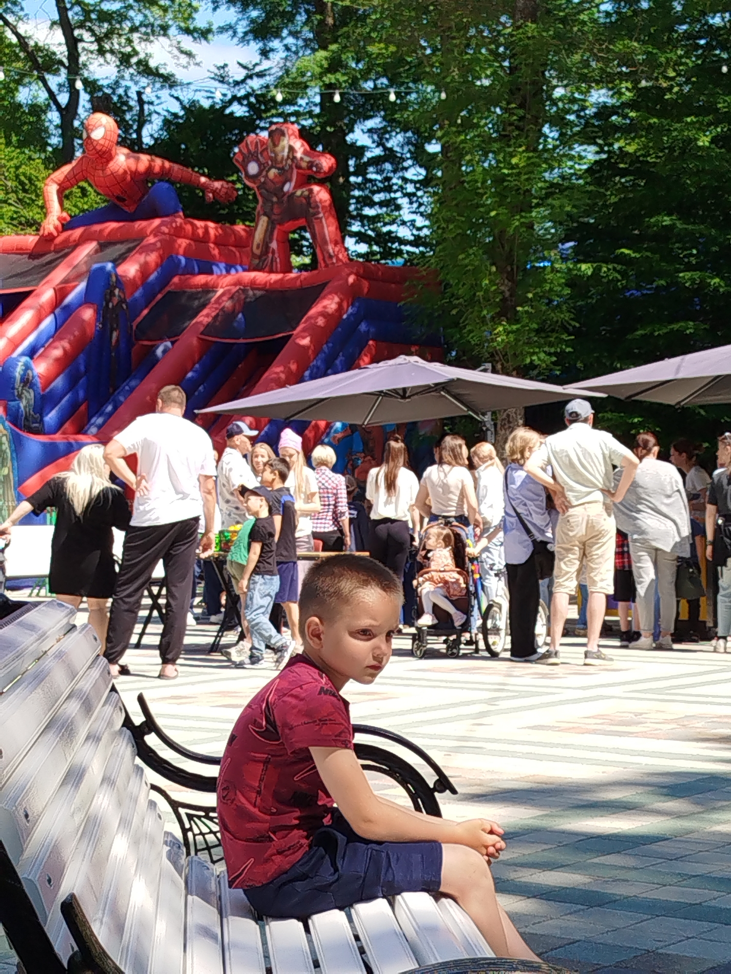 The image depicts an outdoor gathering at a park or fairgrounds, possibly in Stavropol, Russia, on a sunny day. In the background, a large inflatable bouncy castle with Spiderman and Iron Man figures is visible. A crowd of people, including adults and children, are milling around. Some are standing near tables, possibly at a food or game stall, while others are gathered under umbrellas. In the foreground, a young boy with short, light brown hair is sitting on a white park bench, looking off to the side. He is wearing a red t-shirt with a printed design and dark blue shorts. The ground is paved with decorative tiles. Trees with green foliage are present in the background, and some string lights are visible overhead.