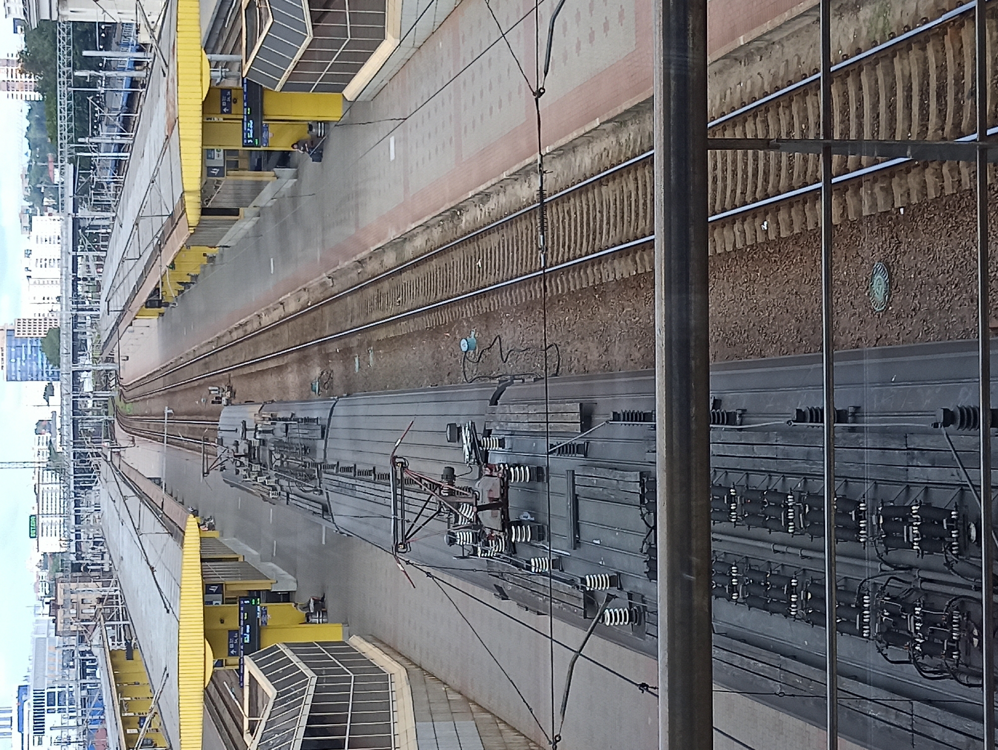 This high-angle, full shot image captures a train station platform in Minsk, Belarus. In the foreground, the top of a train is visible, revealing its electrical pantographs and insulators. The train runs along a track parallel to the platform. The platform itself is covered by a bright yellow canopy supported by pillars. Several digital displays are mounted on the pillars, showing train information. A single person is seated on a bench beneath the canopy, with luggage nearby. The platform is paved with light-colored tiles and bordered by red tiles. In the background, multiple sets of train tracks extend into the distance, surrounded by the urban landscape of Minsk, characterized by buildings of varying heights, including high-rise structures. The sky is overcast. The scene depicts a functional train station environment with elements of the city's architecture visible.