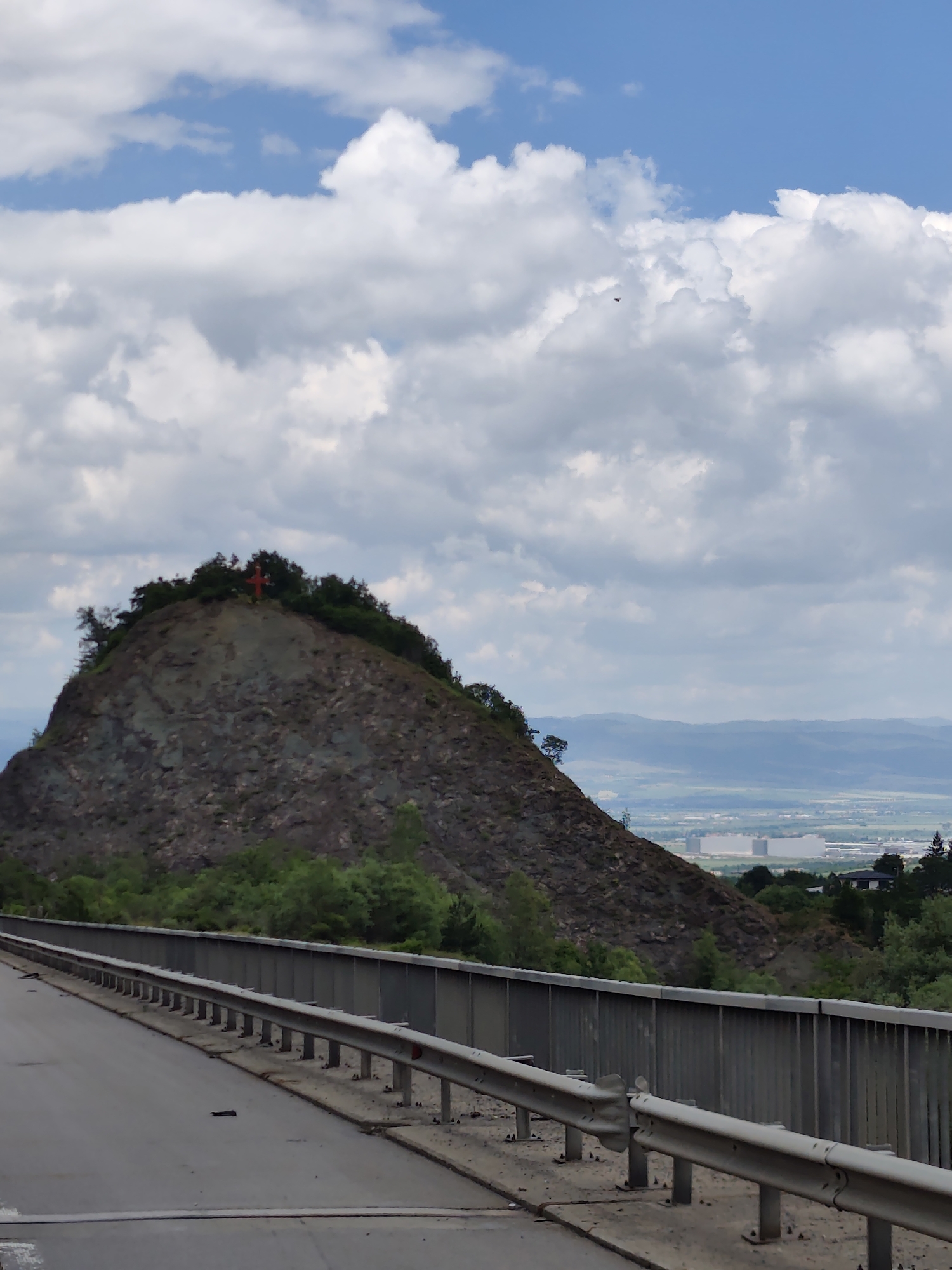 An overcast sky with white and grey clouds hangs above a rugged, tree-covered hill. At the very peak of the hill, a bright red cross is visible, appearing to be a religious monument. The hill is situated in Malo Buchino, Bulgaria. In the foreground, a road with a metal guardrail stretches towards the left side of the frame. Beyond the hill, a hazy landscape unfolds, revealing distant mountains and a sprawling urban or industrial area with large, light-colored buildings. The overall scene suggests a view from a road overlooking a town and its natural surroundings.