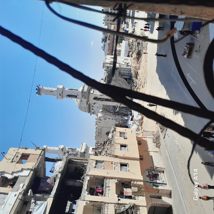 The image depicts a scene of extensive urban destruction in Khan Yunis, Palestinian Territory, viewed from an elevated perspective, partially obscured by dark metal bars or grating in the foreground.
Under a clear blue sky, a heavily damaged multi-story residential building occupies the left side of the frame. Its tan-colored facade shows significant structural collapse, particularly on upper floors, with exposed interiors, rebar, and hanging fabrics visible. Balconies are present but damaged. Graffiti, including a "Squid Game" design, is visible on a lower exterior wall.
In the mid-ground, a severely damaged white mosque with two minarets and a central dome is visible. Portions of its structure, including large sections of walls and the dome, are collapsed or missing. Rubble surrounds its base.
The foreground features a paved street, mostly clear of debris, with a white arrow road marking. Piles of construction rubble and dust are visible off the street, particularly around the mosque and between the structures.
Approximately 10-15 individuals are present on the street and in the background. In the lower left, three children and two adults stand on the street/sidewalk. Further back, individuals are seen walking or standing, including one near a small, makeshift market stall or display of goods on the right side of the street.