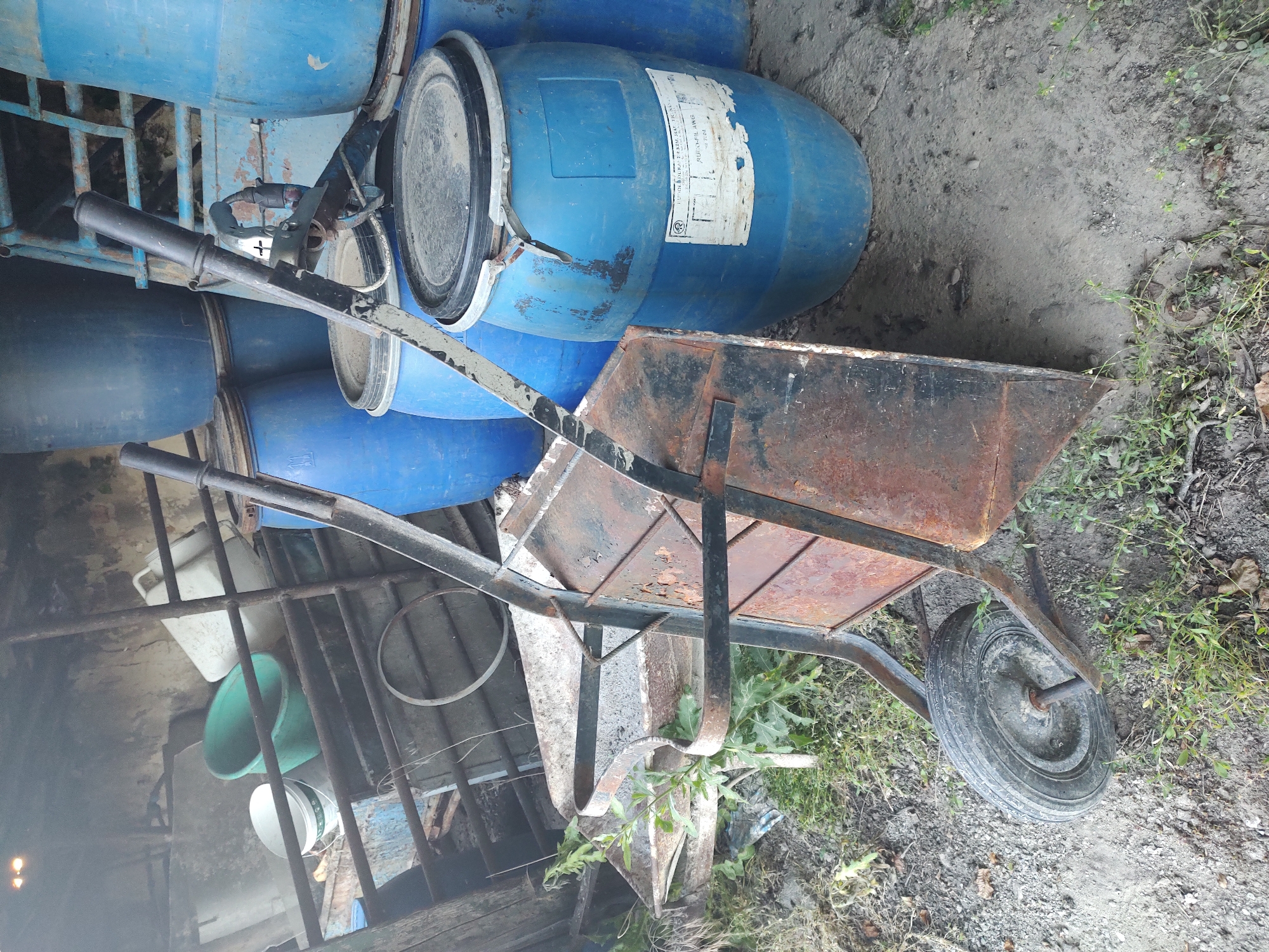 The image shows an outdoor scene, likely in a yard or storage area, with several large blue plastic barrels. In the foreground, a rusted wheelbarrow with a black handle is positioned horizontally across the frame. The wheelbarrow's tray is made of metal and shows signs of wear and tear. A single wheel with a tire is visible on the right side of the wheelbarrow. The ground is a mixture of dirt, gravel, and some sparse green vegetation.
Behind the wheelbarrow, several of the blue barrels are stacked and leaning against each other. One barrel has a white label with some text on it, but the text is mostly illegible. The barrels appear to be storage containers, possibly for liquids. To the left and behind the barrels, there are some metal bars, suggesting a structure like a fence or shelving. Further in the background, a glimpse of a brick wall and more barrels can be seen, along with some green and white containers. The overall impression is one of a storage or work area, possibly a bit unkempt.
There are no people visible in the image. The lighting suggests it could be daytime, possibly overcast given the lack of strong shadows. The scene appears to be located in a rural or semi-rural setting, given the ground cover and the presence of the barrels and wheelbarrow, which are commonly found in such environments. The visible text on the barrel label is too blurry to be read.