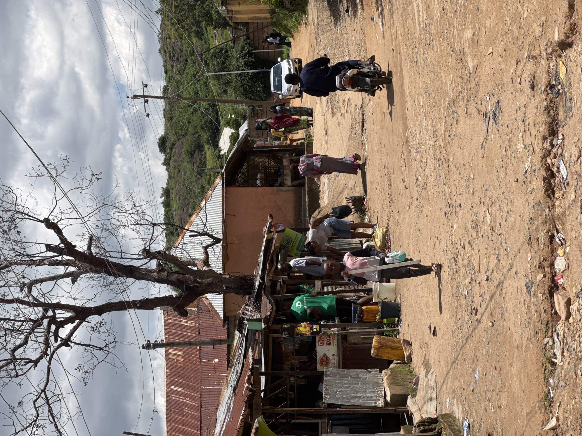 The image depicts a dusty, unpaved street in a neighborhood in Jos, Nigeria. A young boy, dressed in a pink and white shirt and dark pants, walks down the middle of the street carrying a white object. To his left, a makeshift stall or shop with a corrugated metal roof displays various goods. Several adults are visible around the shop, including a man in a green t-shirt. In the background, a white car is parked on the street, and a person is riding a motorcycle. The street is lined with modest buildings, some with corrugated metal roofs, and lush green hills rise in the distance under a cloudy sky. The ground is littered with debris and has several potholes.