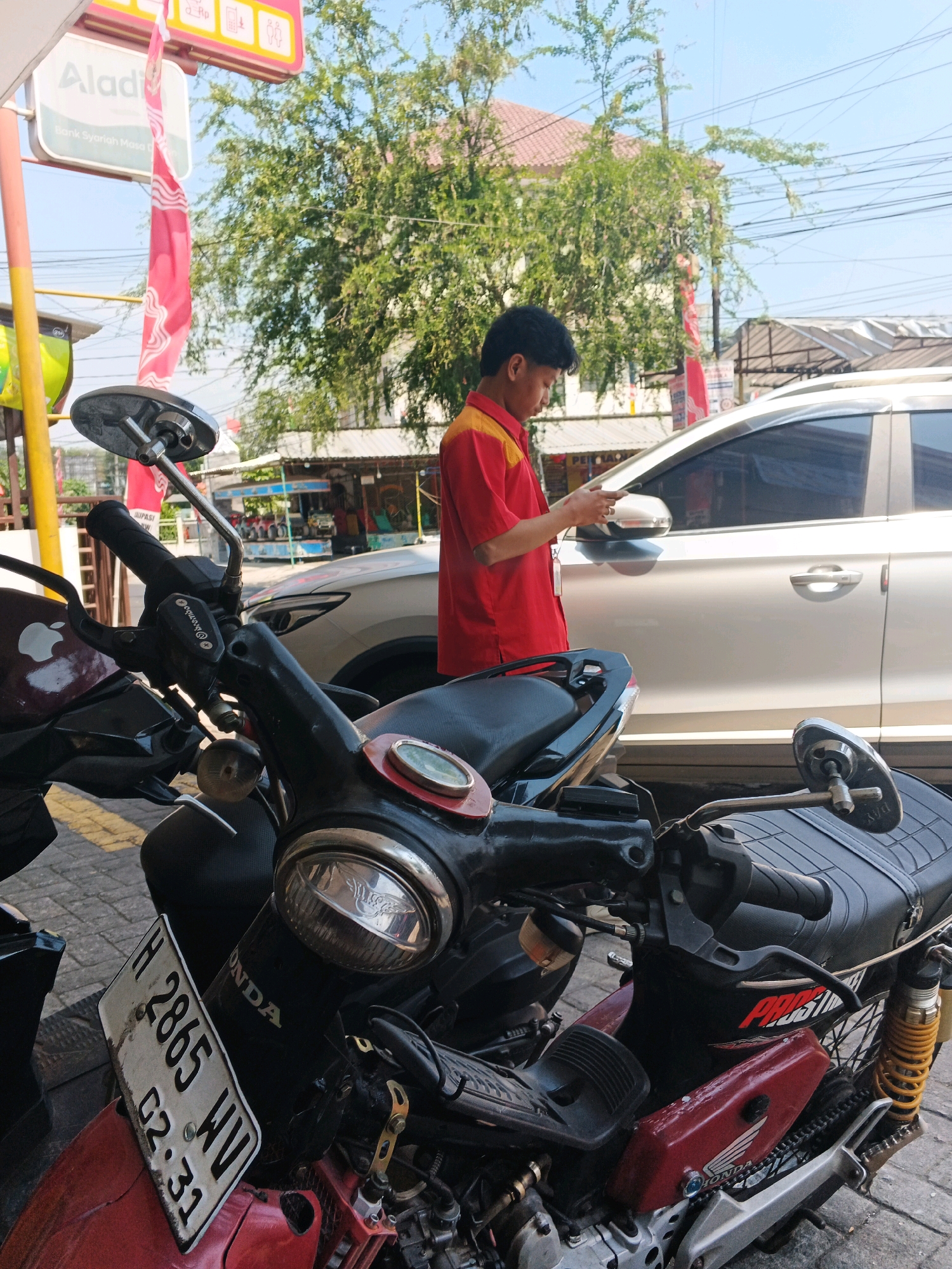 A young man in a red and yellow shirt is standing next to a silver car, looking at his phone. In the foreground, a red and black motorcycle is parked. The scene appears to be in a city, possibly in Kalirejo, Indonesia, given the Indonesian license plate on the motorcycle and the presence of a store with Indonesian signage. The background shows buildings, trees, and power lines, suggesting an urban or semi-urban environment. The lighting indicates it is daytime.