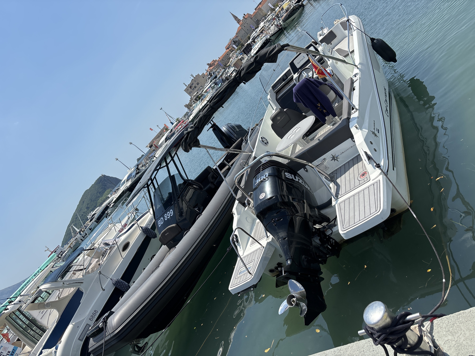 The media file depicts a scene of several boats docked in a harbor, with a town and hills in the background. The boats appear to be a mix of motorboats and possibly a rigid-hulled inflatable boat. One of the motorboats prominently features a Suzuki outboard motor. There is visible text on the boats, including "GARMIN" on a GPS unit, registration numbers like "BD 899", and model names such as "Cap Camar" and "YHW-SURF". The scene is set under a clear blue sky, suggesting daytime with fair weather. The boats are moored next to a concrete dock, with mooring lines visible. The overall impression is one of a recreational boating area, likely in a coastal town. The buildings in the background, characteristic of European architecture, combined with the geographical features like the hills, help to identify the location as Budva, Montenegro.