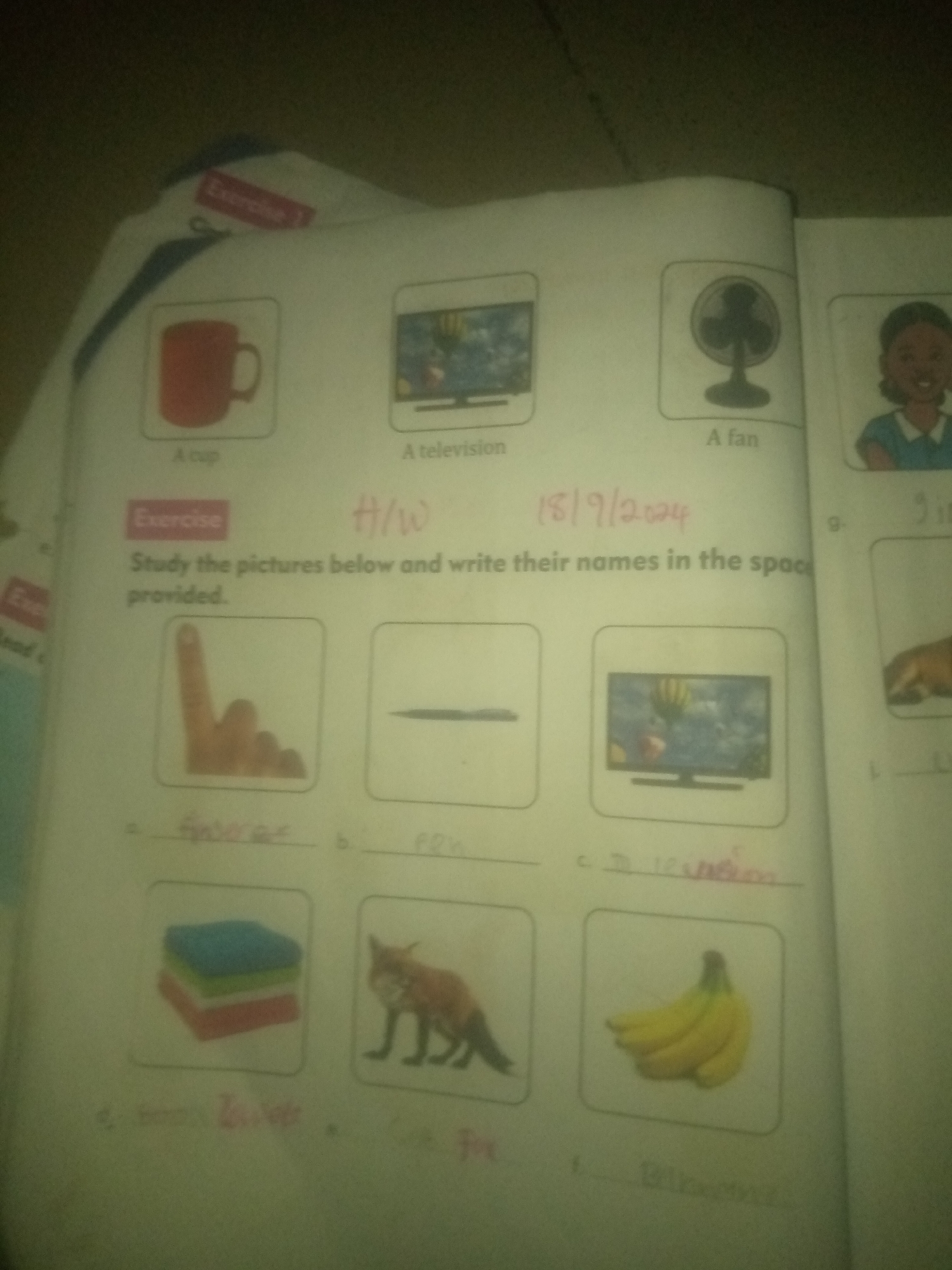 The image is a photograph of an open book, likely a children's workbook, on a flat surface. The book has a pink "Exercise" label and handwritten "H/W" and a date "18/9/2024". The workbook contains images of various objects and animals, each with a space for a written name.
In the top row, there are pictures of a red cup, a television, a fan, and a girl. Below these are boxes with:
a) A hand with one finger raised, with the handwritten word "finger" underneath.
b) A pencil or pen, with the handwritten word "pen" underneath.
c) A television, with the handwritten word "Television" underneath.
d) A stack of colorful folded clothes, with the handwritten word "Clothes" underneath.
e) A fox, with the handwritten word "Fox" underneath.
f) A bunch of bananas, with the handwritten word "Bananas" underneath.
The image does not contain any information about the location in Jauro Yinu, Nigeria. It is a close-up shot of the book, with the background being dark and out of focus.