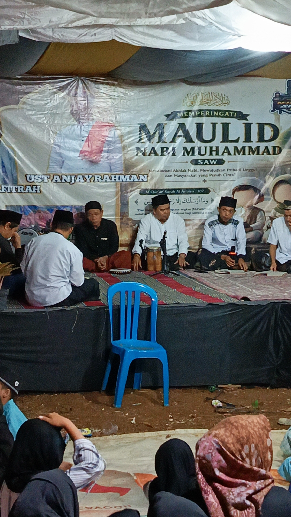 In Margajaya, Indonesia, an outdoor event is taking place, likely a religious gathering. A large banner with Islamic calligraphy and text is hung, featuring the words "MAULID NABI MUHAMMAD SAW" and "UST ANJAY RAHMAN". The banner also includes Quranic verses in Arabic.
Several men are seated on a raised platform covered with a patterned cloth. They are dressed in traditional Islamic attire, including kufis (caps) and long-sleeved shirts. Some men are holding microphones, suggesting they are speakers or performers. One man is playing a musical instrument resembling a small drum.
In front of the platform, a bright blue plastic chair stands alone. The ground in front of the platform appears to be covered in dirt and scattered with debris. In the foreground, a group of people, mostly women wearing hijabs, are seated on the ground, facing the platform. Their backs are mostly turned to the camera. The overall scene suggests a community event focused on religious commemoration.