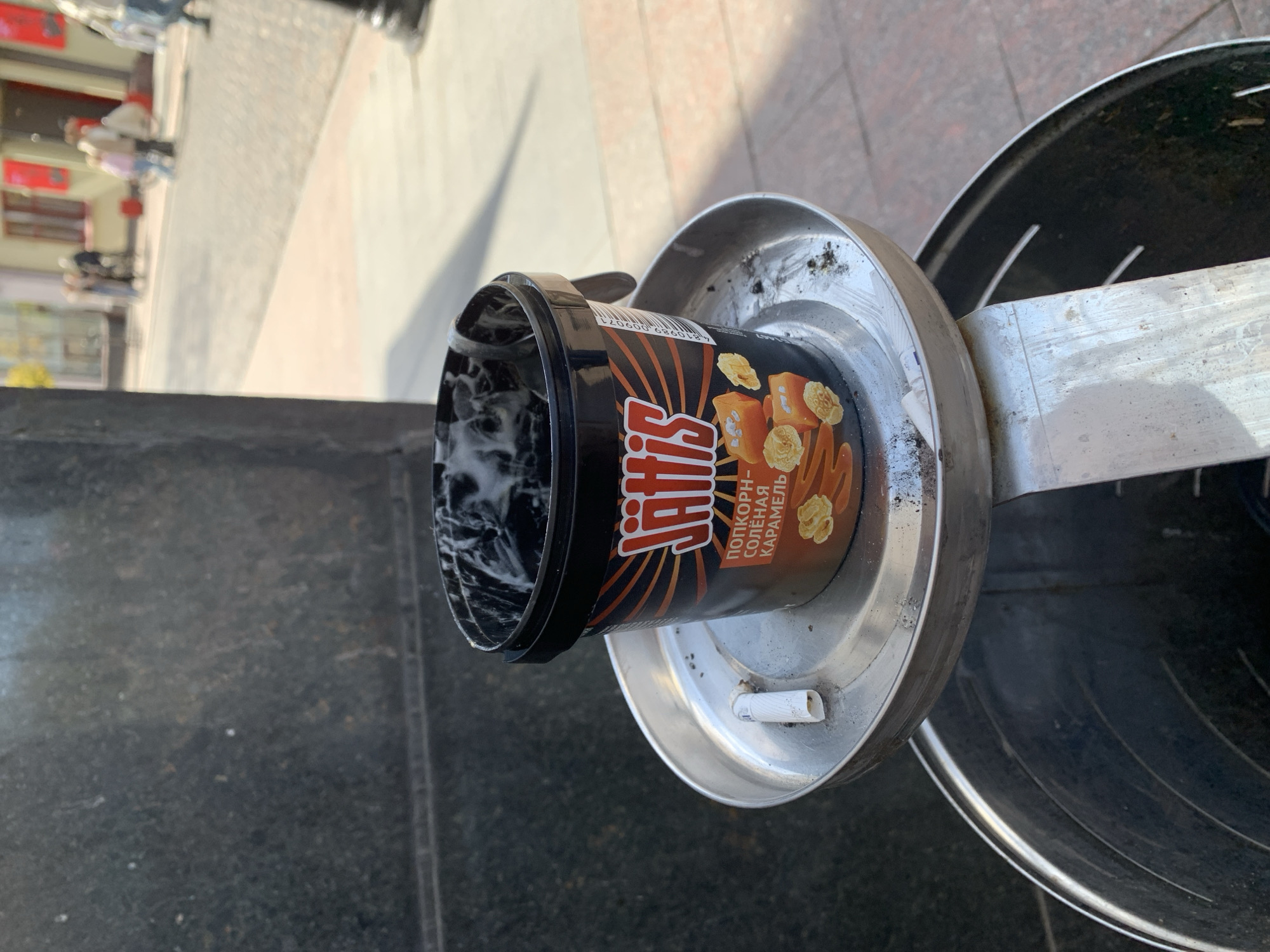 The image depicts an empty Jattis ice cream tub precariously balanced on a metallic ashtray, which is integrated into a public trash can. The setting is an outdoor urban environment, consistent with a street or public square in Hrodna, Belarus.
The ice cream tub is black with orange and brown accents, featuring the brand name "JATTIS" prominently. Below it, Cyrillic text reads "ПОПКОРН-СОЛЁНАЯ КАРАМЕЛЬ" (Popcorn-Salted Caramel), with illustrations of popcorn kernels and caramel squares. A barcode is also visible on the side of the tub. The inside of the tub contains some white residue, possibly melted ice cream or condensation.
The metallic ashtray is circular and shows signs of use, with some dirt and smudges. A discarded white cigarette butt rests in one of its circular depressions. The ashtray is attached to a larger, dark-colored metallic trash can, only partially visible on the right side of the frame. The ground beneath is dark, possibly paved with stone or asphalt tiles.
The background is softly blurred, showing an expanse of light-colored paving, distant buildings with red accents, and indistinct figures of people, suggesting a busy street or pedestrian area. The scene is illuminated by bright natural light, indicating it is daytime. The activity captured is the disposal of a finished ice cream container and a cigarette butt in a public receptacle, reflecting a common urban occurrence.